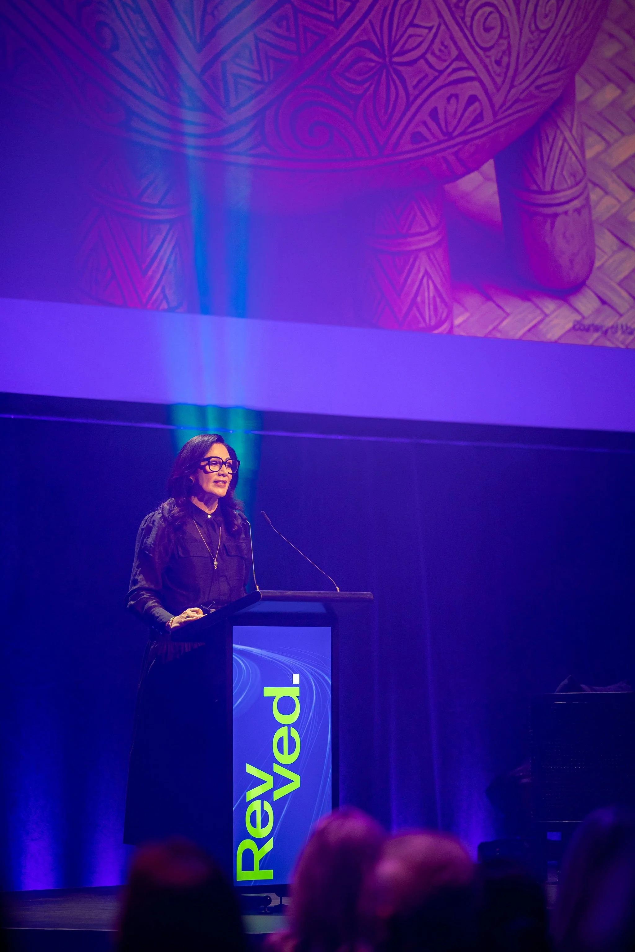 AA speaker presenting at a podium on stage during Revved, with a large screen behind her displaying abstract graphics.