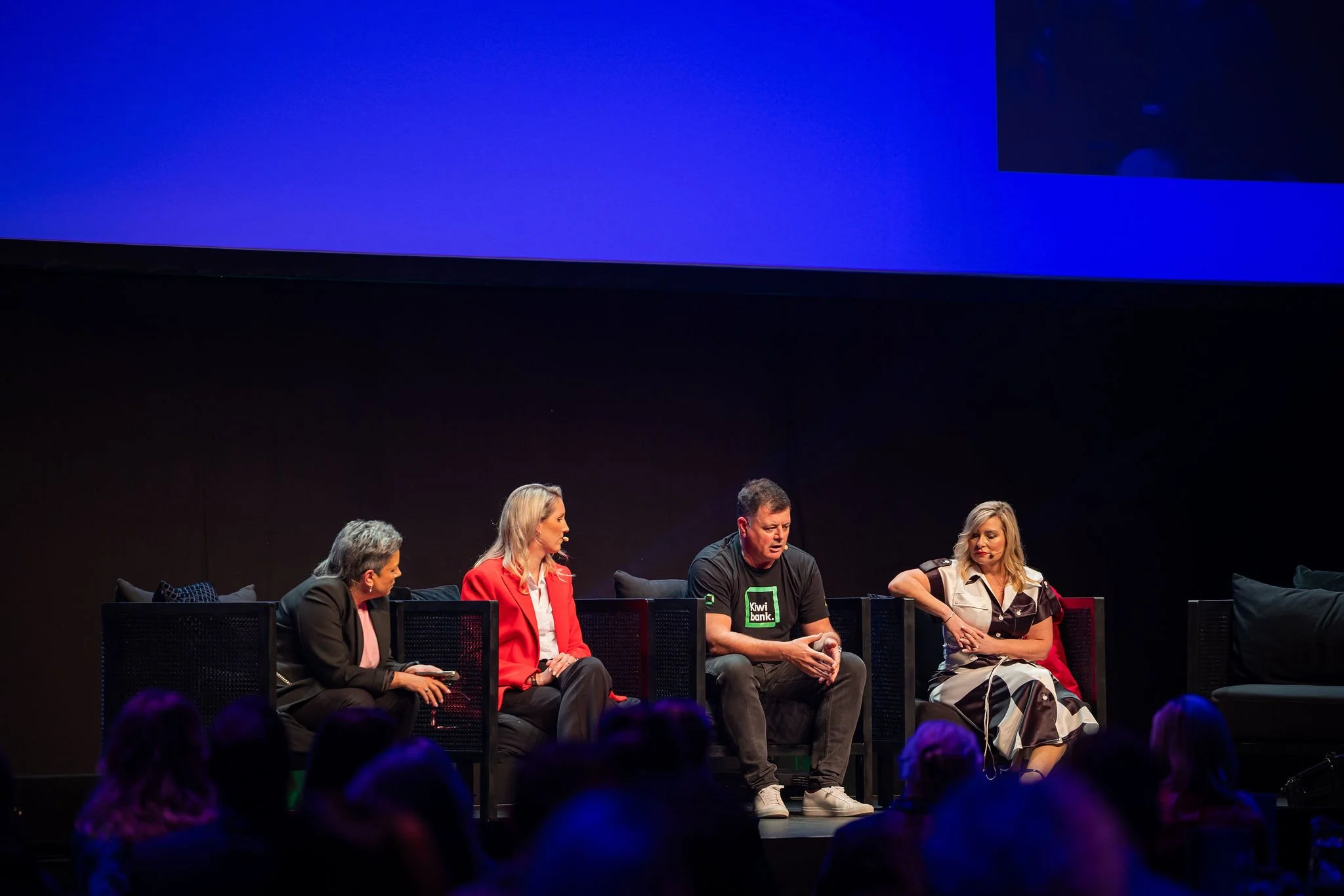 Group of five people sitting on chairs on a stage, engaged in a panel discussion or conference, with a dark background and a large blue screen above them.