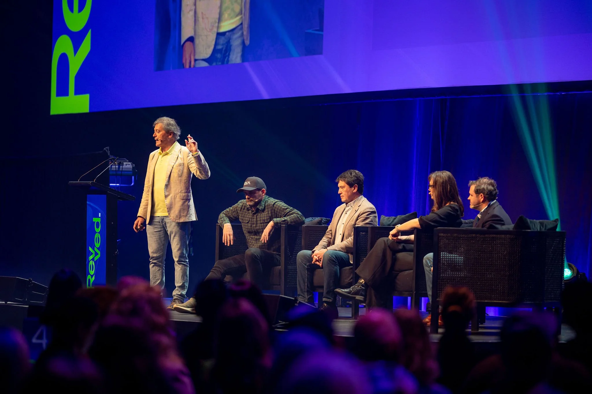 On stage at Revved, with five people, one standing and speaking at a podium, and four seated in a row behind them, during a conference or panel discussion, with a large screen and colorful lighting.