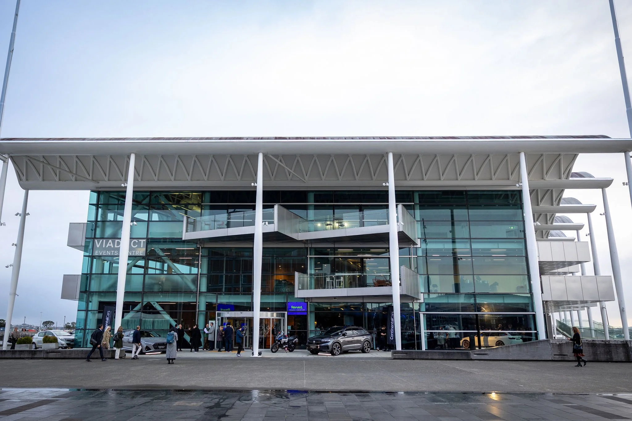 Modern building with glass facade and a large overhanging roof, labeled as an events center named 'VIADUCT.' Several cars are parked outside, and people are walking near the entrance on a cloudy day.