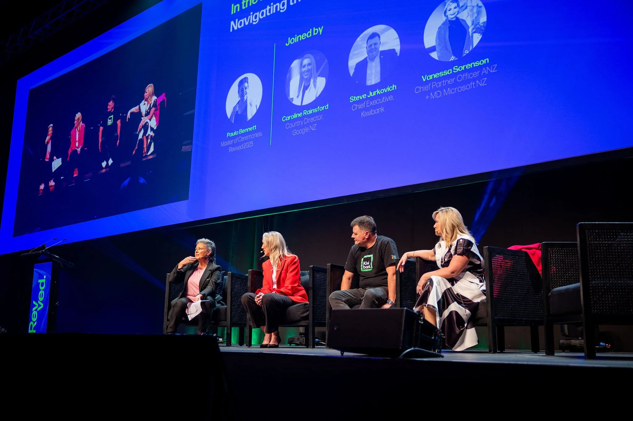 A panel of women seated on stage at a conference with a large screen behind them displaying profiles and names of speakers, including Caroline Rainford, Steve Jurkovich, and Vanessa Sorenson. The audience is visible in the foreground.