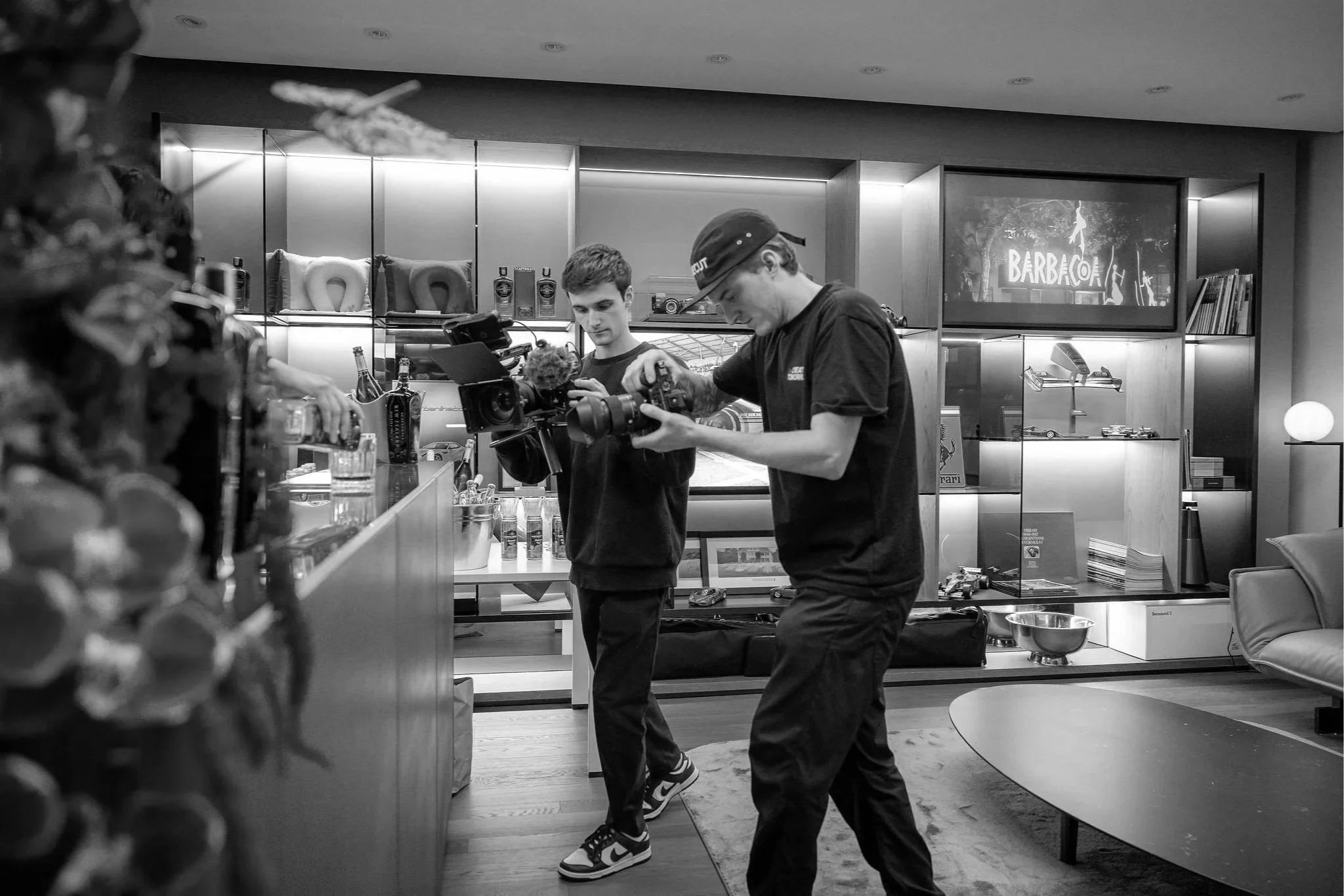 Two young men working with a professional video camera in a modern living room or studio, with shelves displaying brands and a large screen showing the word 'Barboca' in the background.