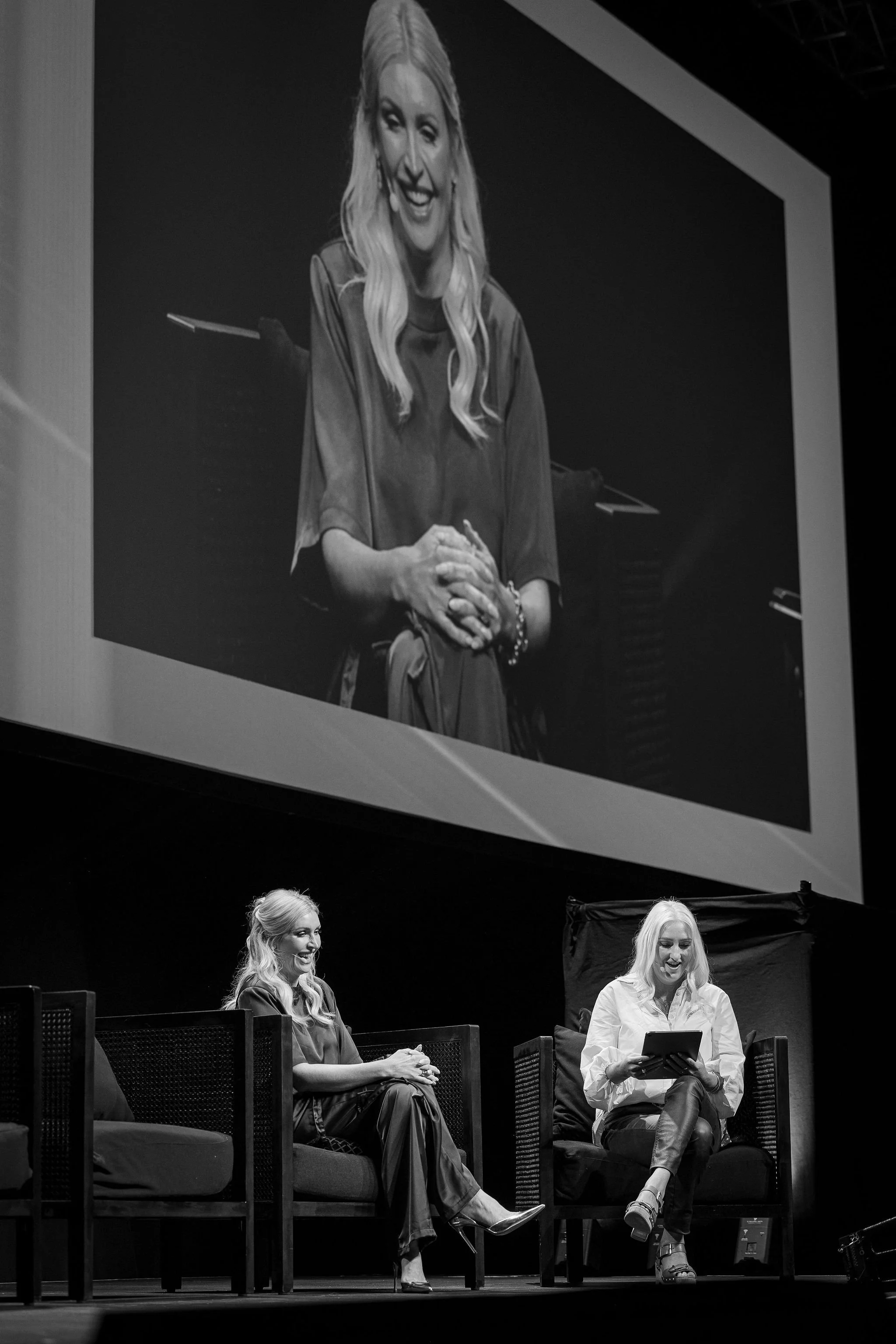 Black and white photo of two women sitting on stage, with a large screen behind them displaying a woman smiling. One woman on the right is holding a tablet, and the woman on the left appears to be laughing.