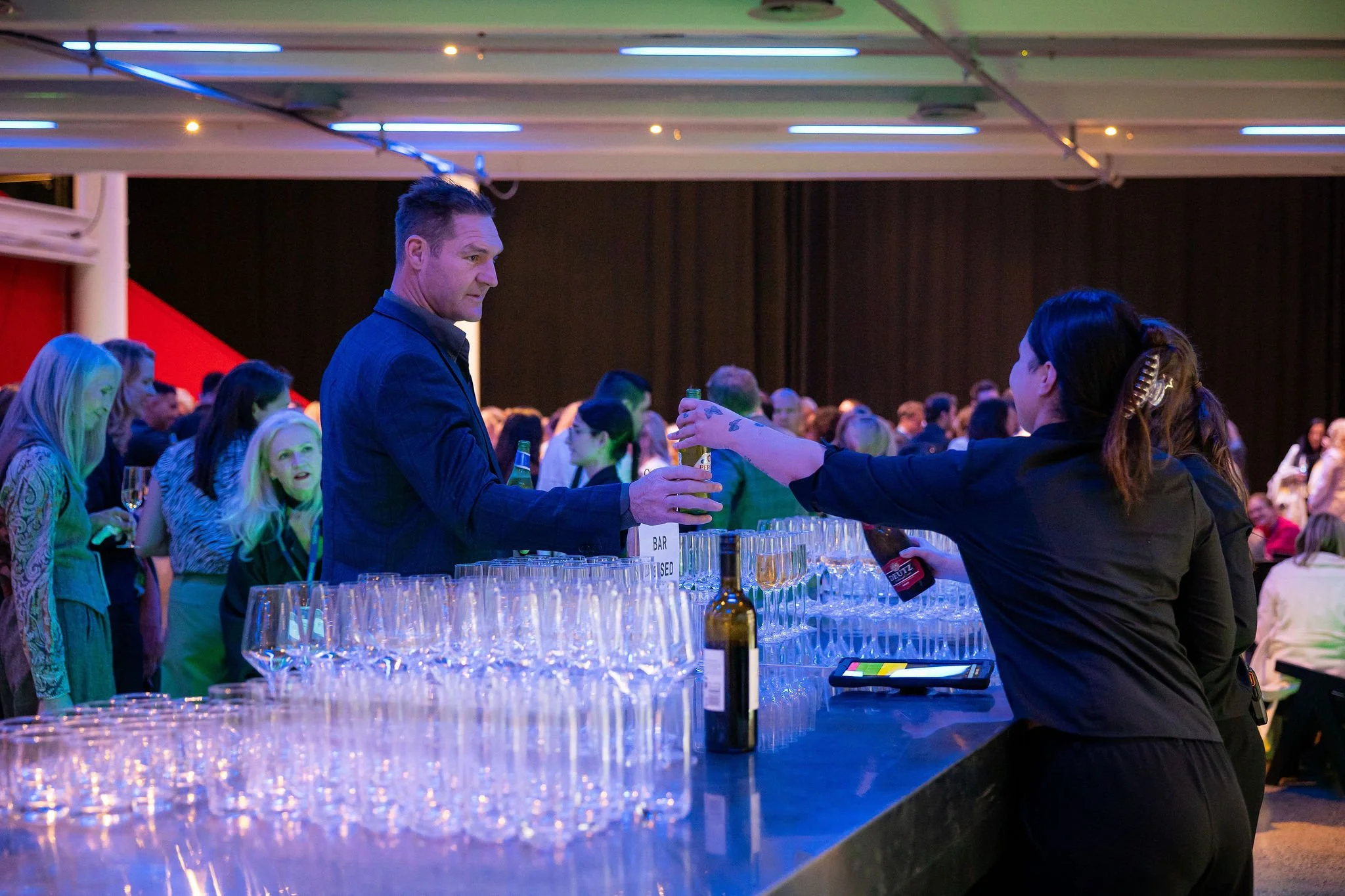 At a busy social event, a man in a dark suit is holding a beer bottle and talking to a woman behind a bar. The bar is set with many empty glasses and bottles. People are gathered in the background, mingling and enjoying the event, with colorful light
