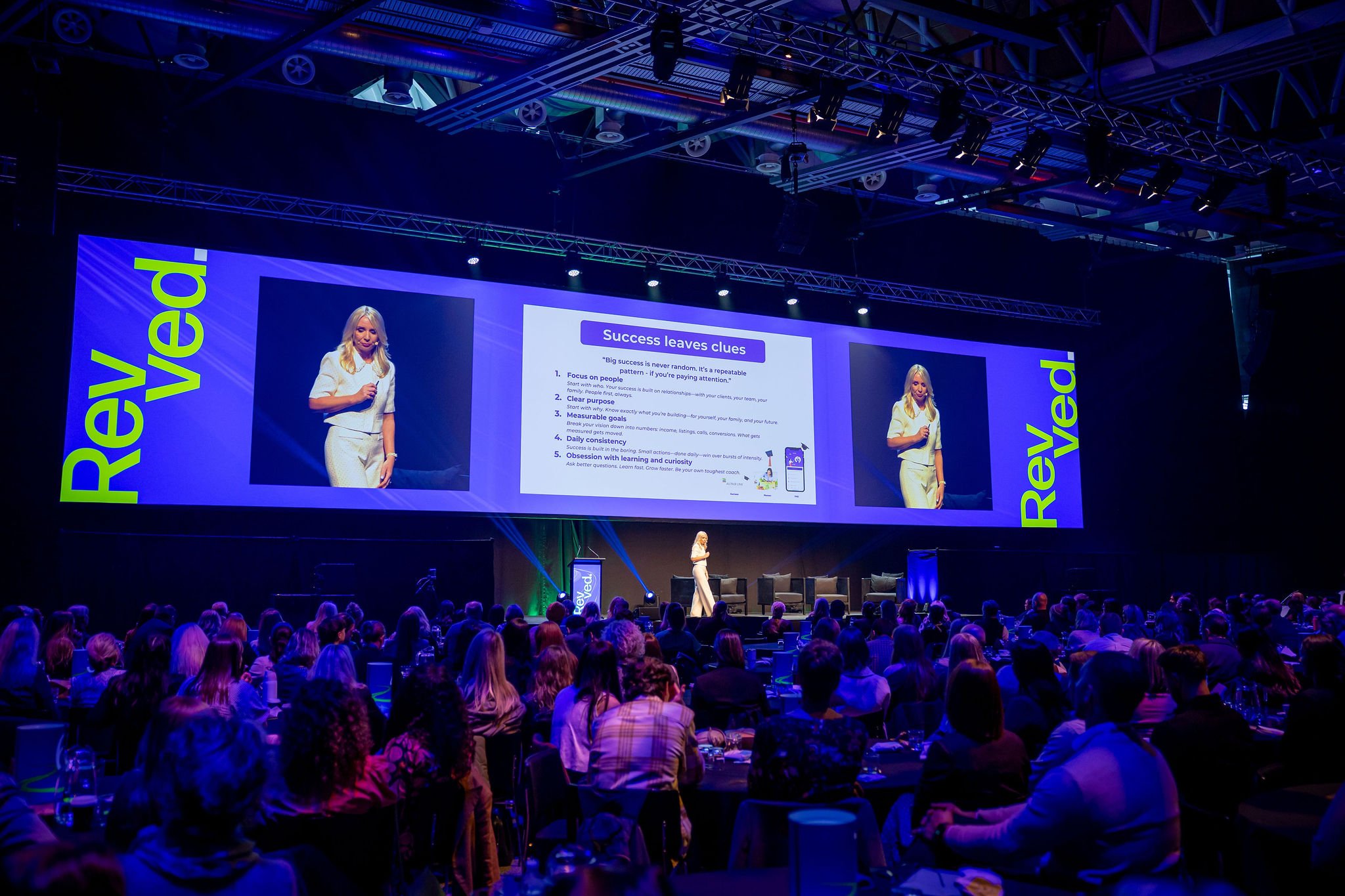 A large audience attending a conference or seminar with a woman presenter on stage. The stage has a large screen displaying presentation slides titled 'Success leaves clues' with tips. The woman is dressed in white and stands at a podium.
