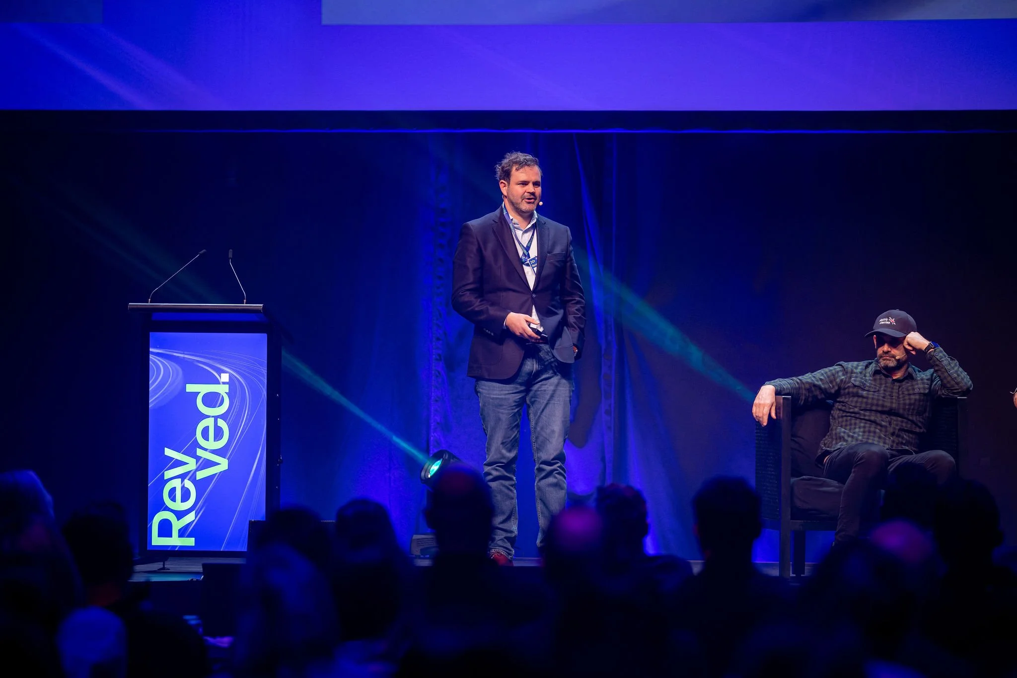 A presenter at Revved with a large screen behind him displaying the word "Revved" and blue lighting. 