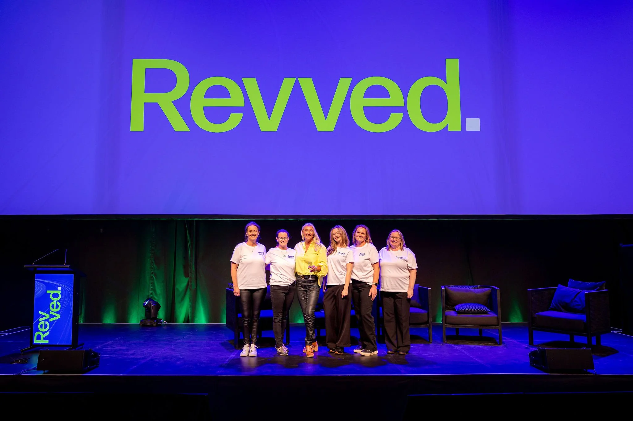 Group of six women standing on stage during a presentation or event, with a large screen behind them displaying the word 'Revved' in green font on a blue background. The women are smiling and wearing casual white T-shirts.