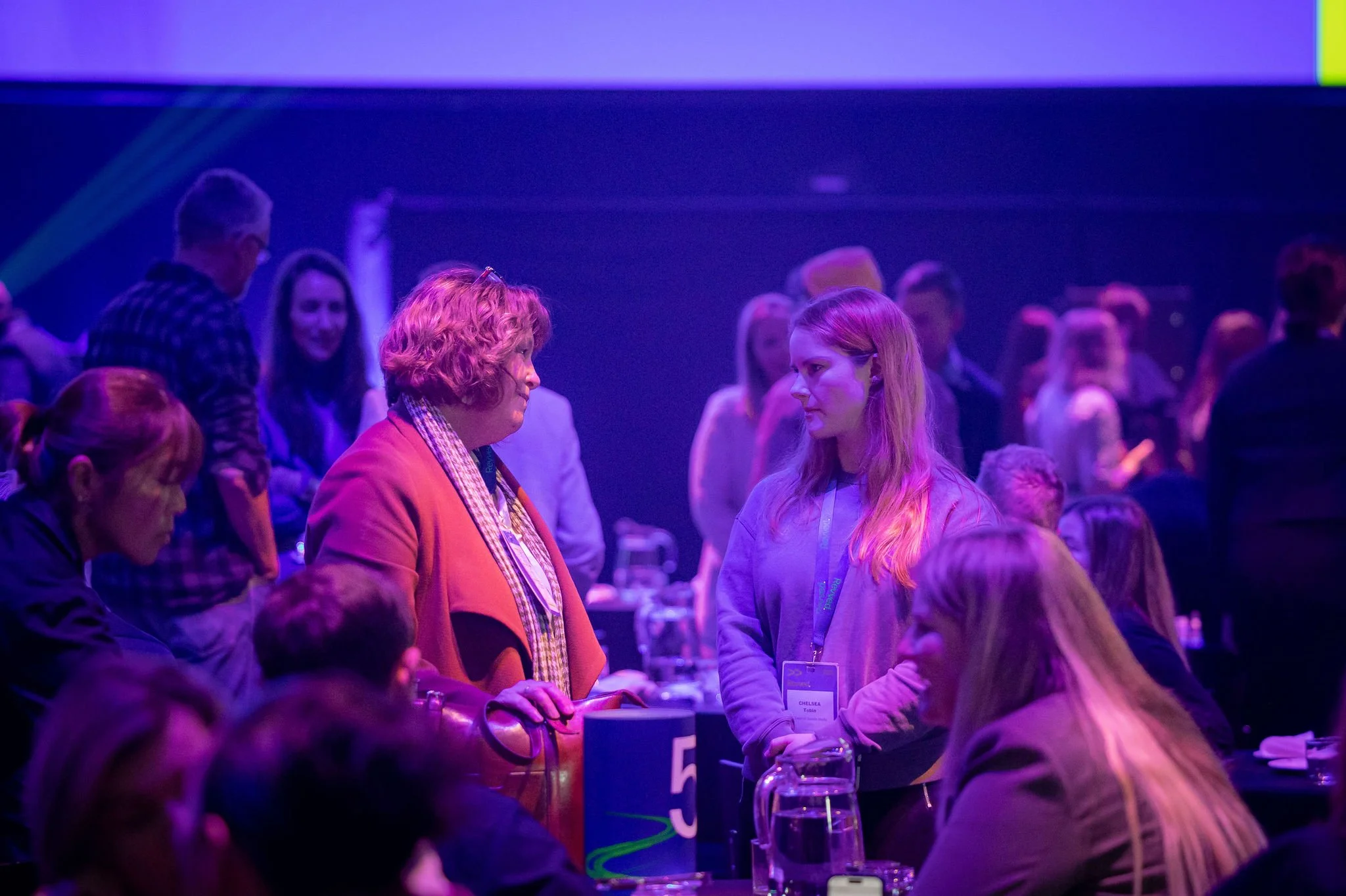 Group of people gathered at a conference or event, engaging in conversation, with some wearing name tags, in a dimly lit room with purple lighting.