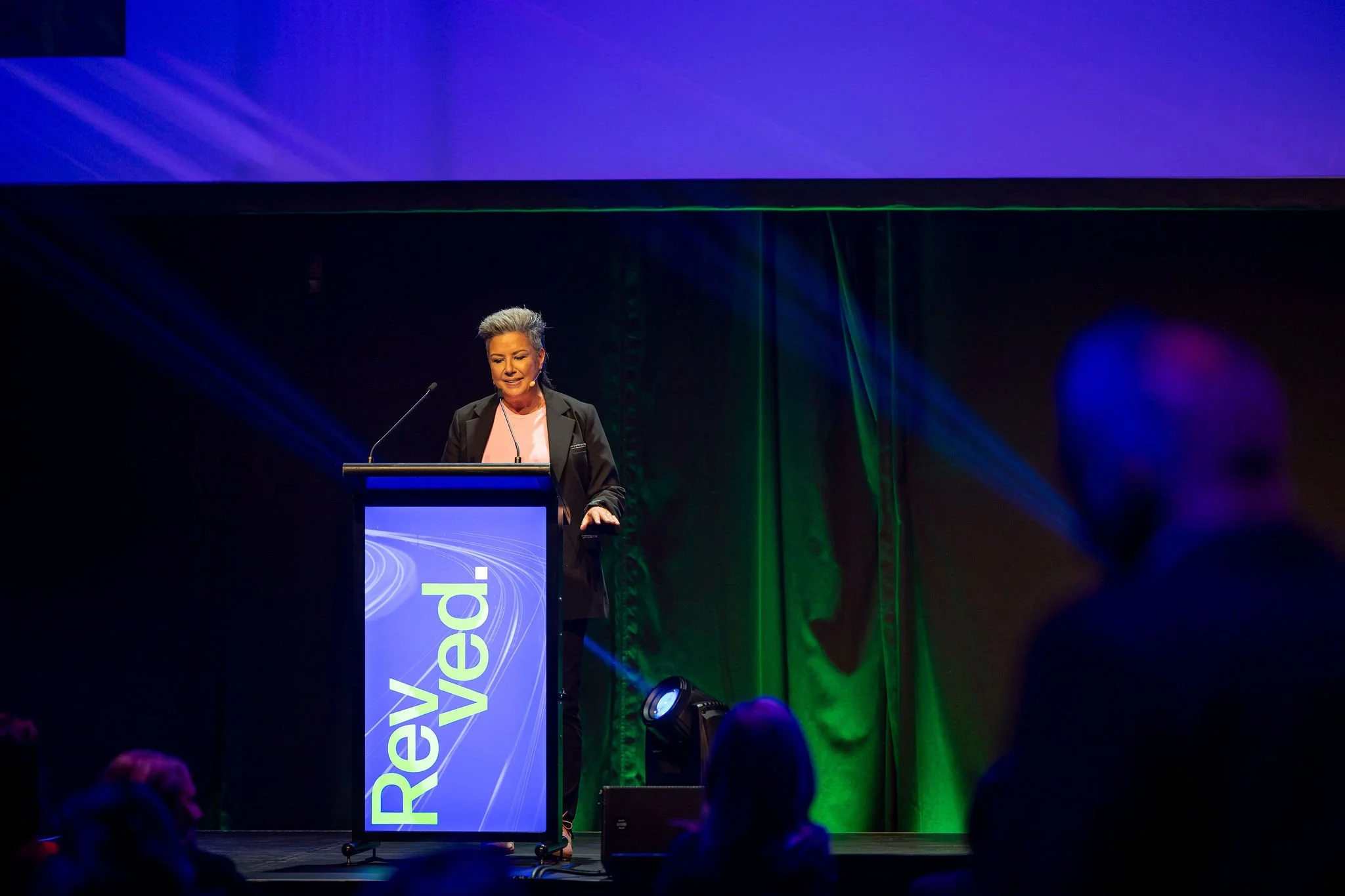 A woman speaking at a podium on a stage with a large screen behind her displaying the word 'Revolution' in bright colors, with audience members visible in the foreground.