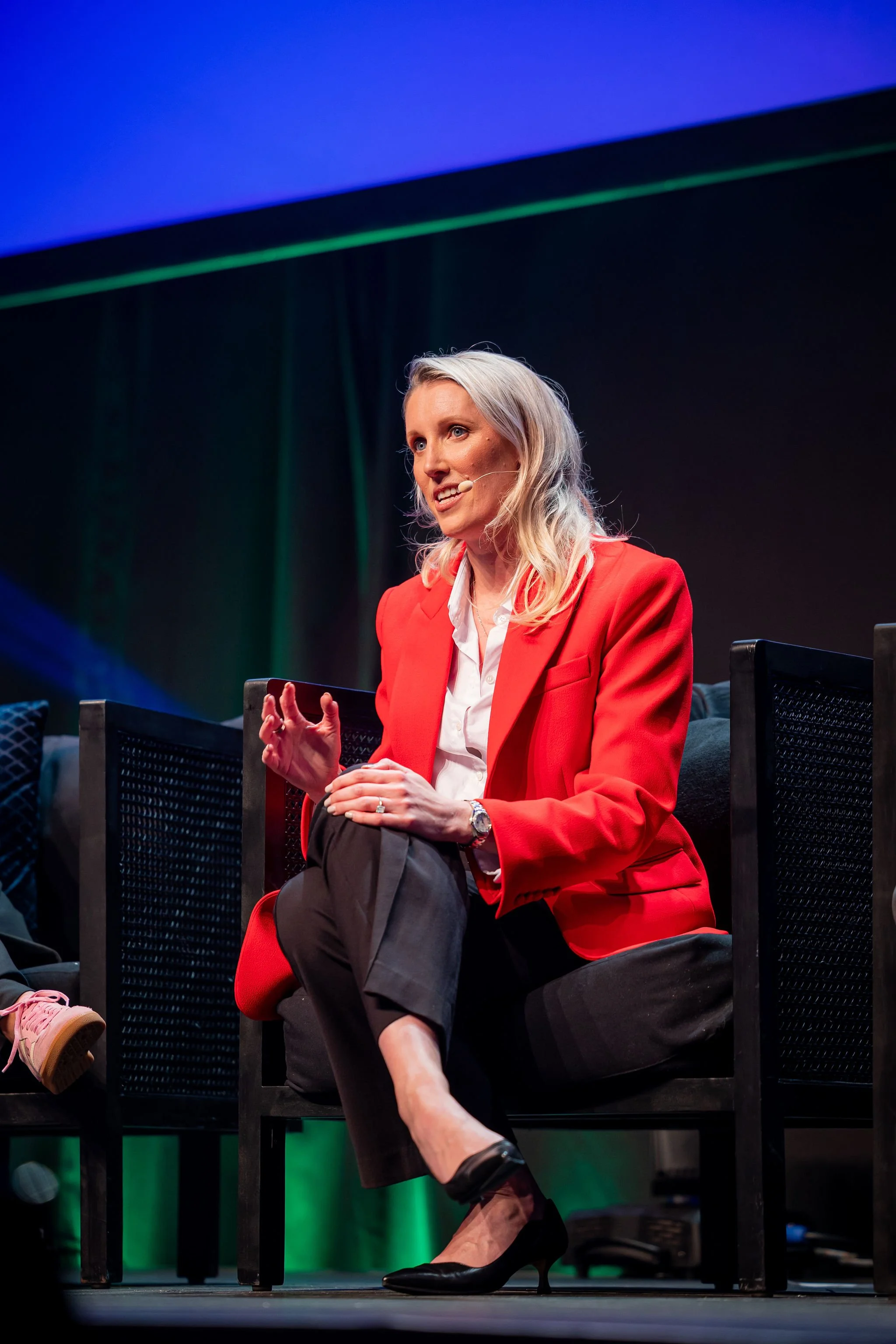 A woman in a red blazer speaking on a stage, sitting on a black chair with her legs crossed, wearing black high heels, with a dark background and colorful stage lighting.