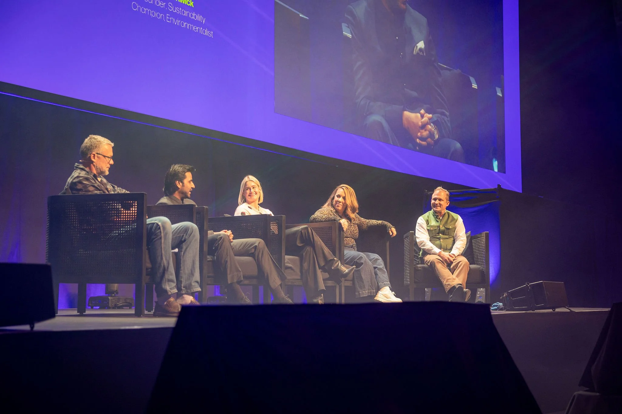Revved conference panel discussion on stage with six people sitting in chairs, large screen behind them displaying a mirrored image of the stage, with blue lighting and a dark background.