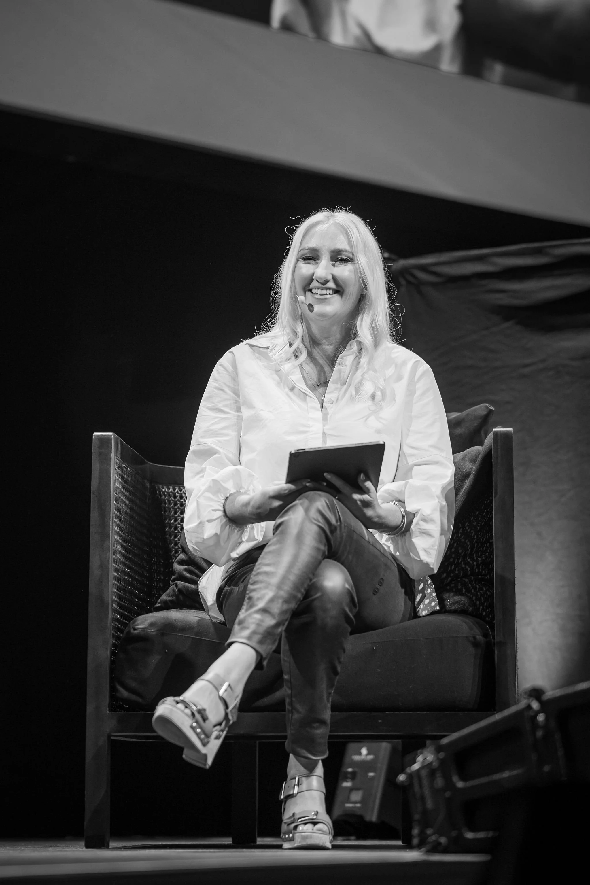 A woman with long blonde hair smiling, sitting on a chair on stage, holding a tablet, wearing a white shirt and pants, with her legs crossed, on a dark stage with a large dark backdrop.