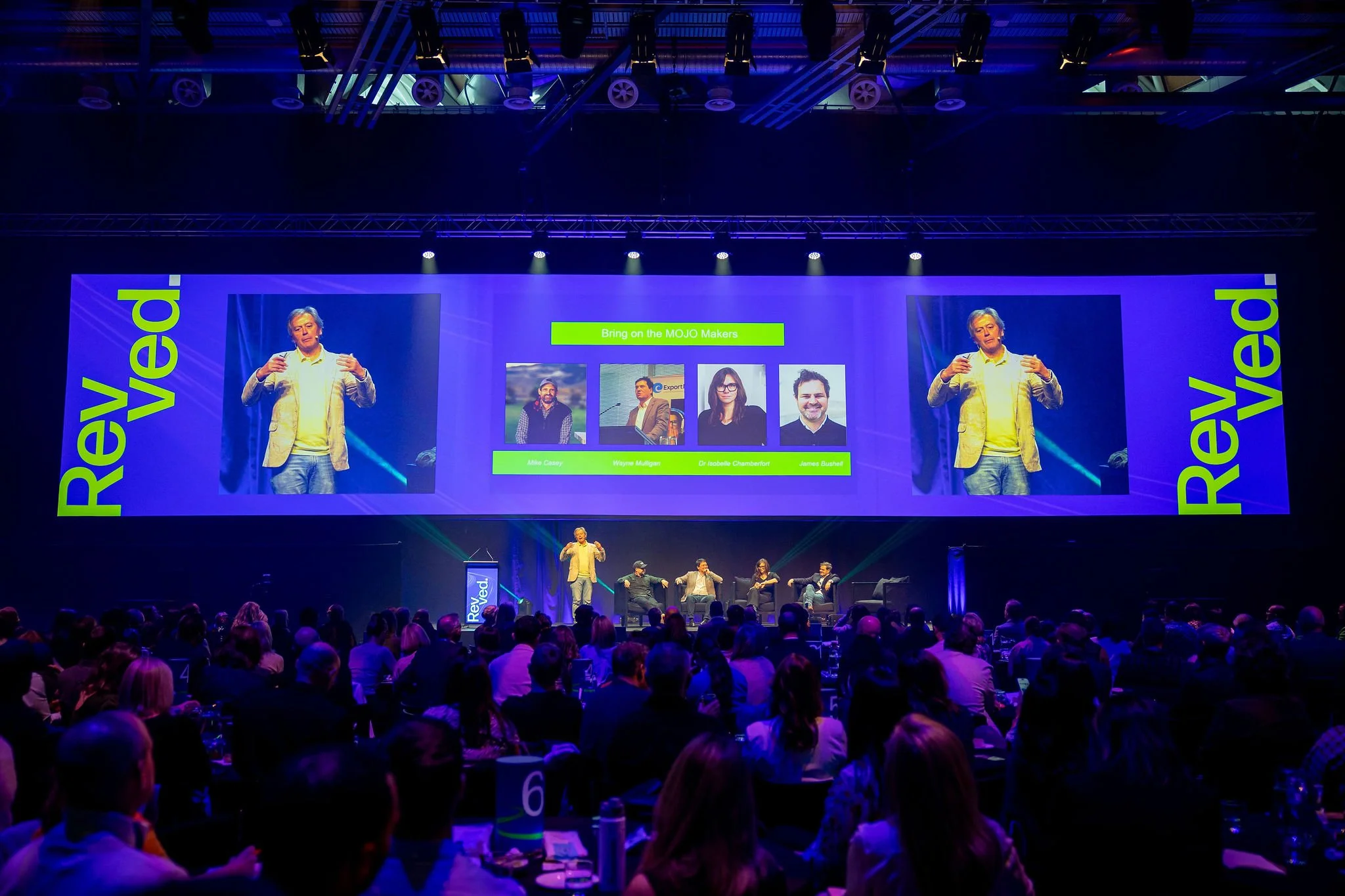 A stage at Revved with speakers and a large audience, featuring a screen with four panelists' photos and names, and the words "Revved." The speakers are engaged, with one standing and gesturing.