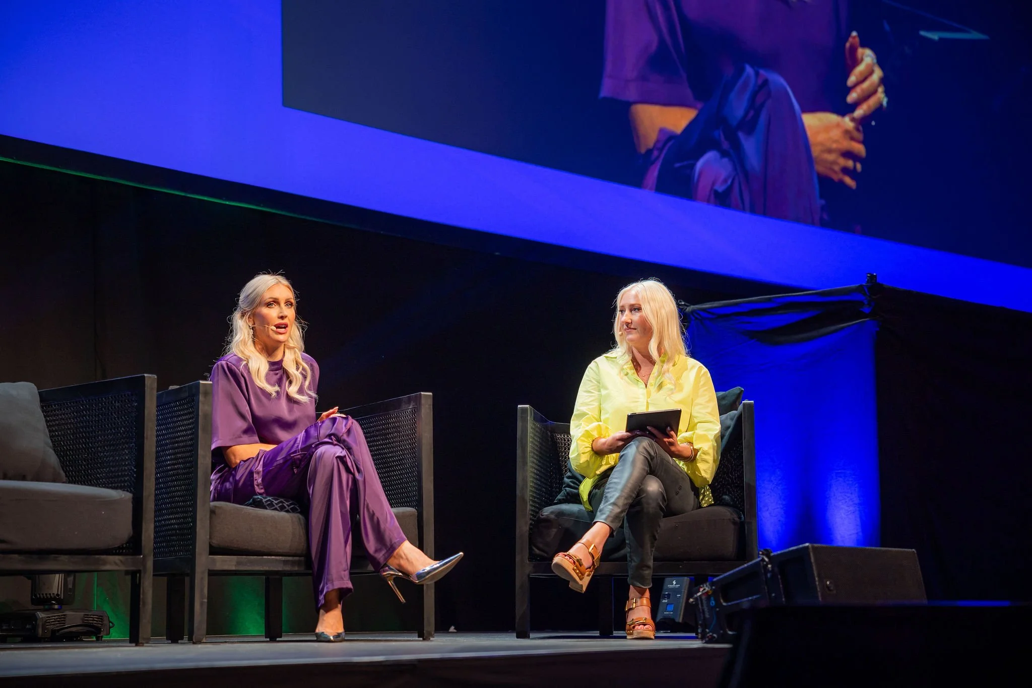 Two women sitting on stage during a panel discussion, one in purple and the other in yellow, with a large screen behind them projecting their image.