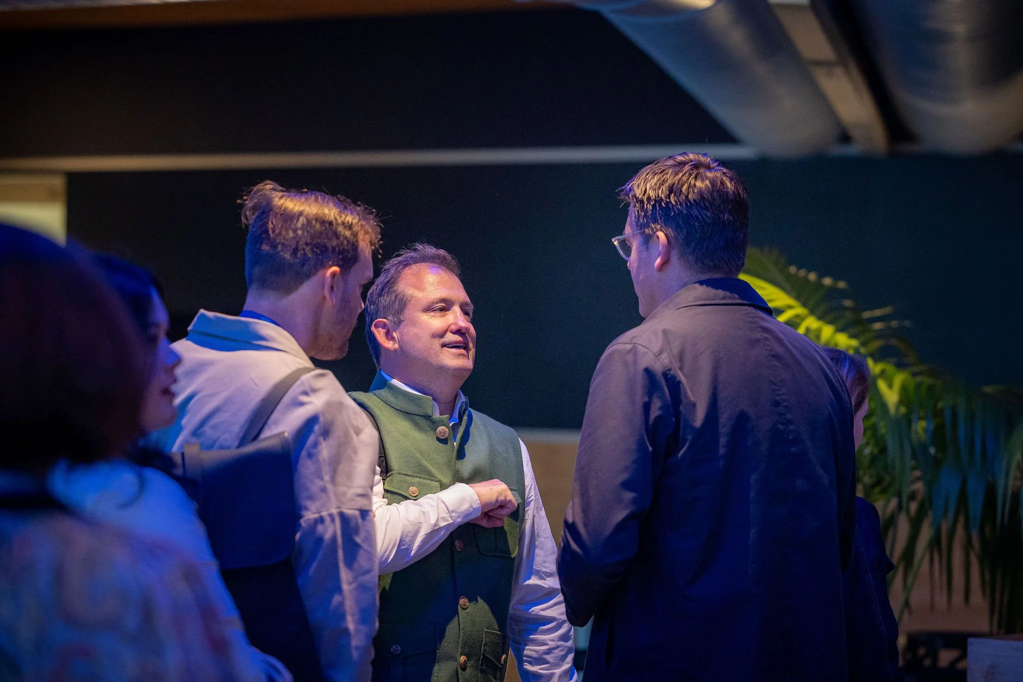 Group of four men in conversation, one wearing glasses, dark background, indoor setting with plants.
