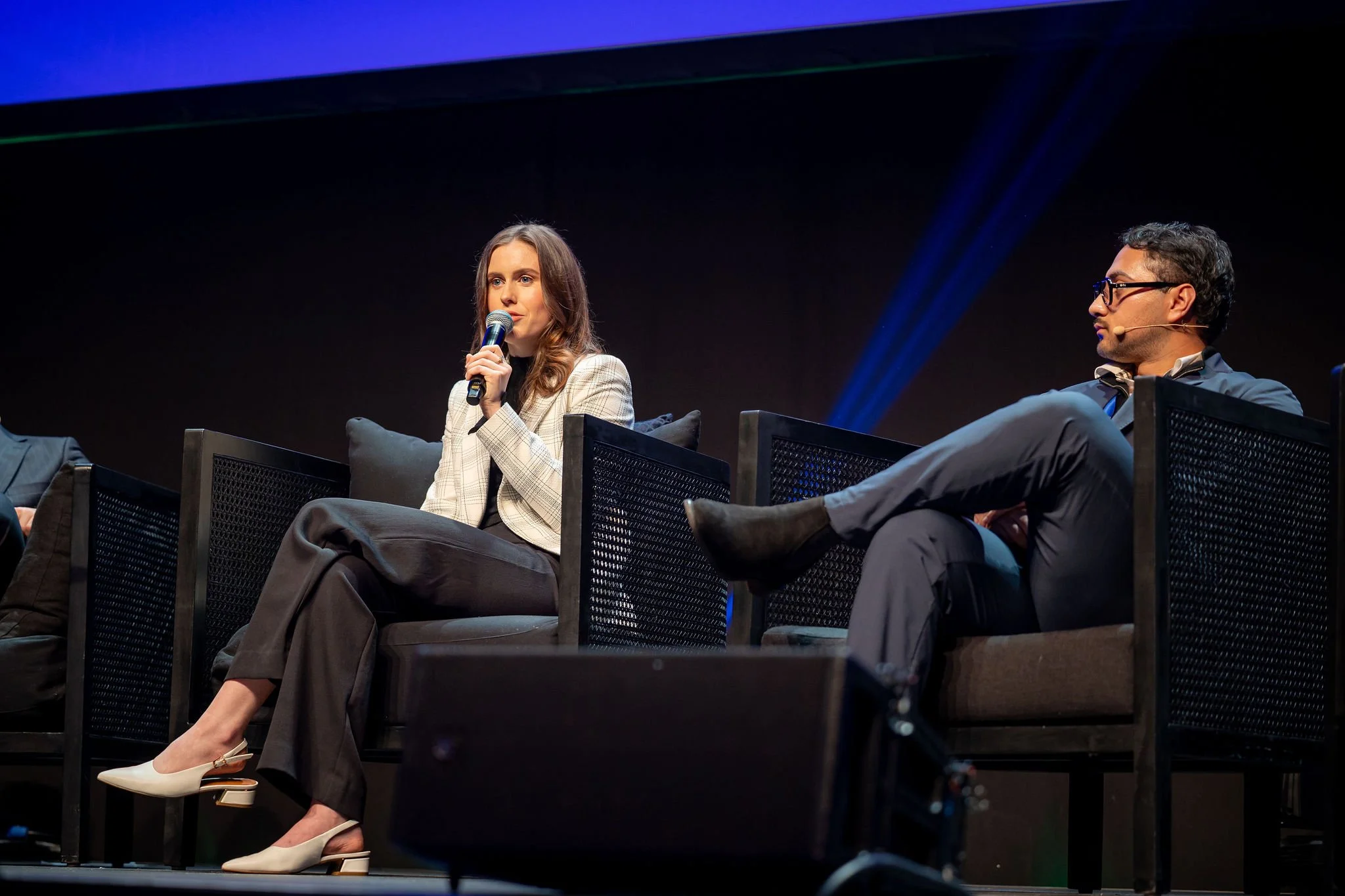 A woman speaking into a microphone on stage during a panel discussion, sitting on a black chair, wearing a plaid blazer, black pants, and beige heels, with a man wearing glasses and a suit sitting next to her.