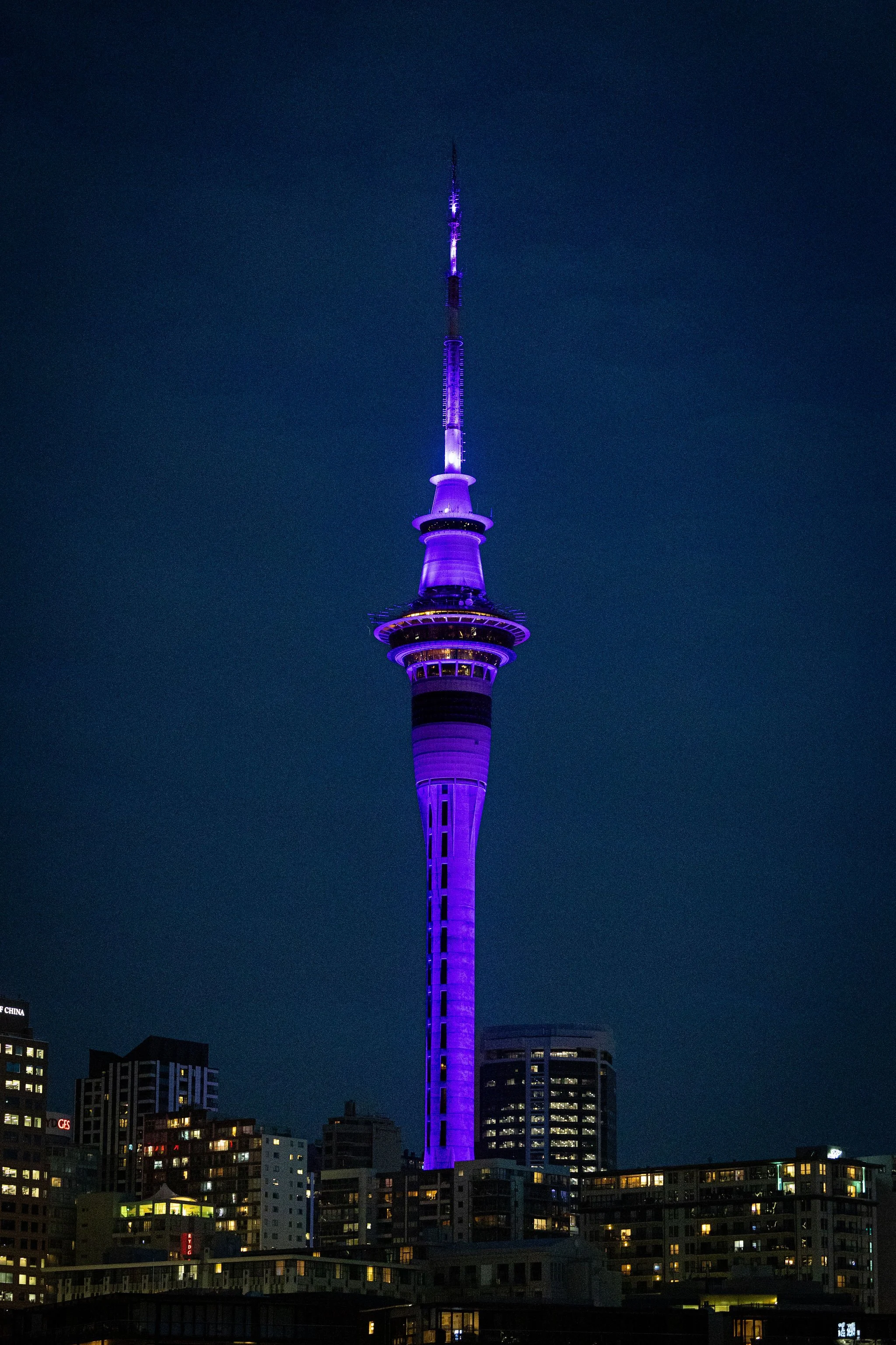 A tall skyscraper tower illuminated in purple lights against a dark night sky, with surrounding city buildings with lit windows at the base.