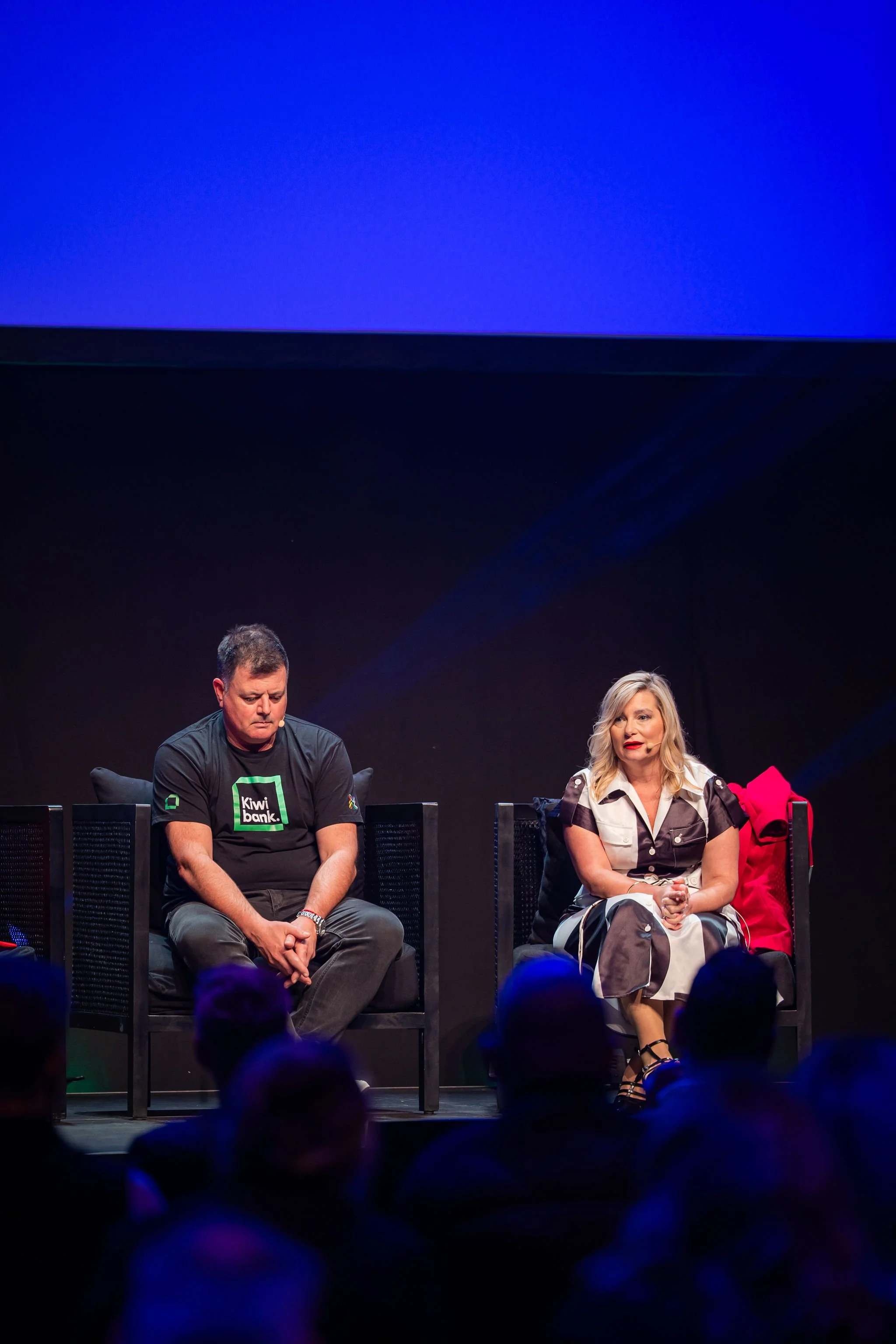Two people sitting on stage during a panel discussion, with a blue screen in the background and audience members visible in the foreground.