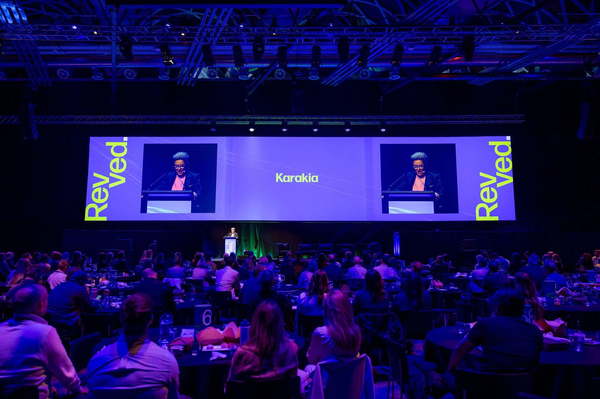 A large conference hall filled with attendees sitting at round tables, listening to a speaker on stage. The stage has a large screen displaying the name 'Karakia' and the words 'Revived' on side panels. The room is lit with blue and purple lighting, 