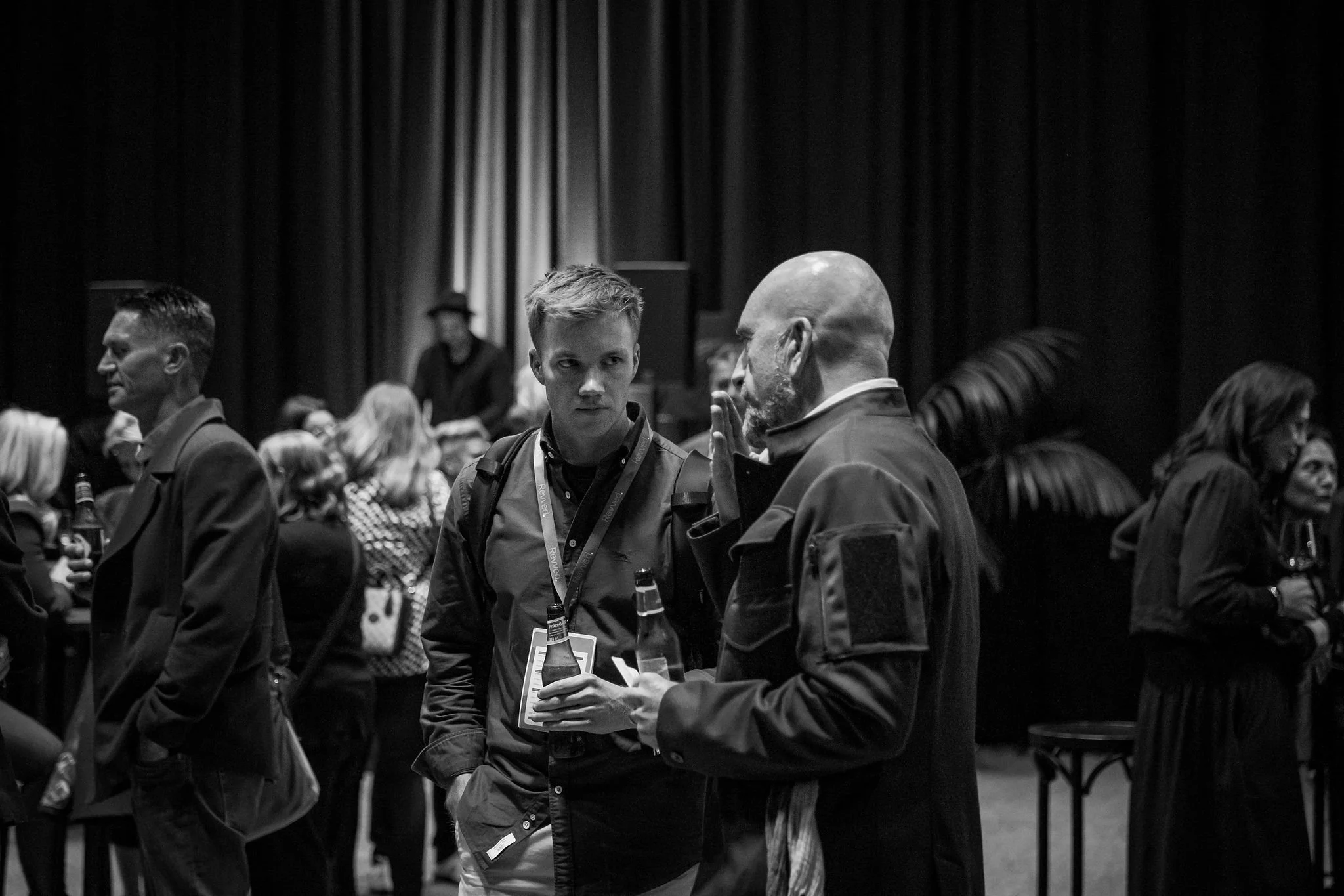 Black and white photo of people at a conference or event, with two men in the foreground engaged in a conversation, surrounded by seated attendees.