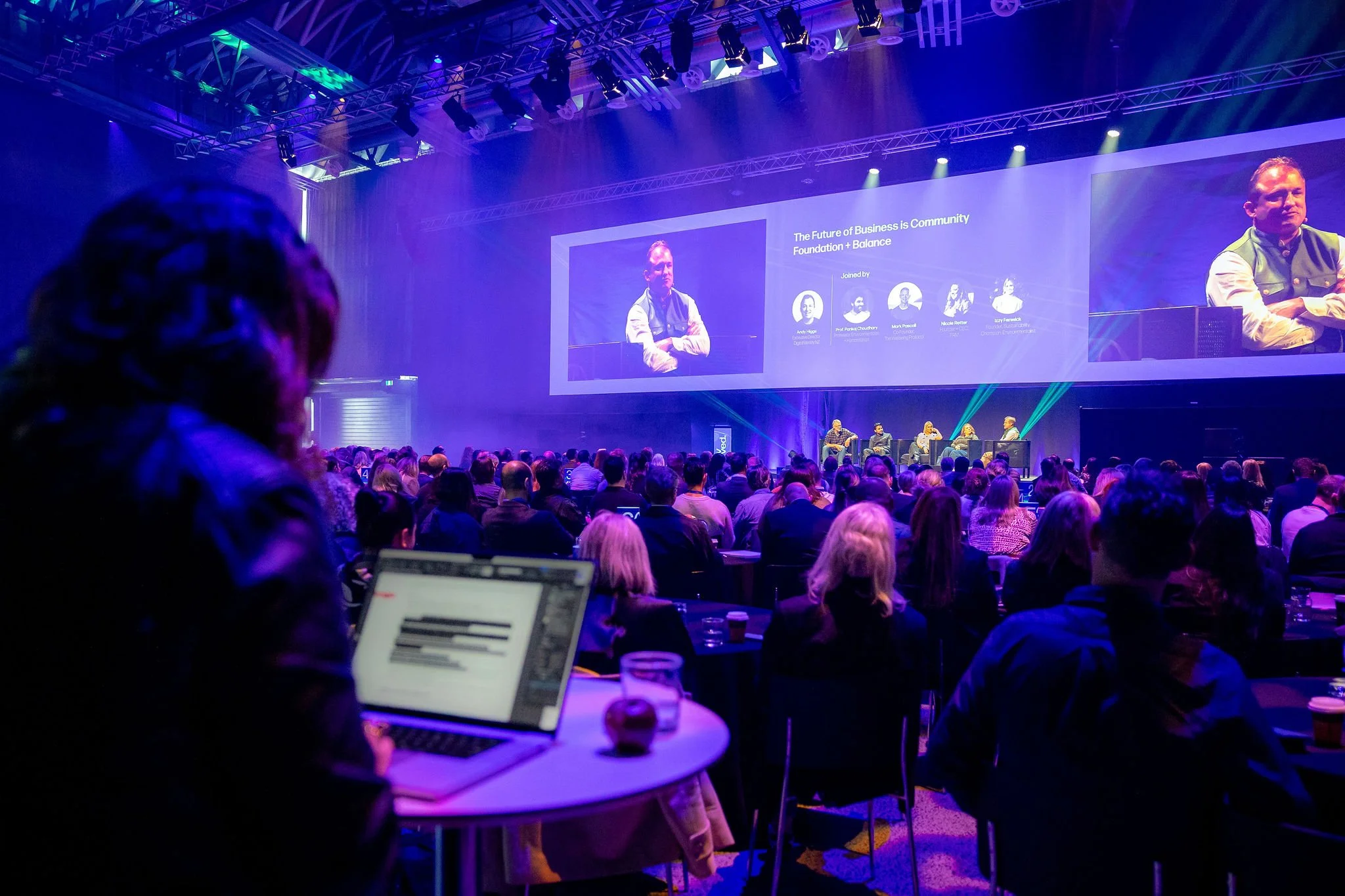 Revved conference with many attendees seated facing a stage. The stage has a large screen displaying panelists and presentation information. Some audience members are using laptops or devices.
