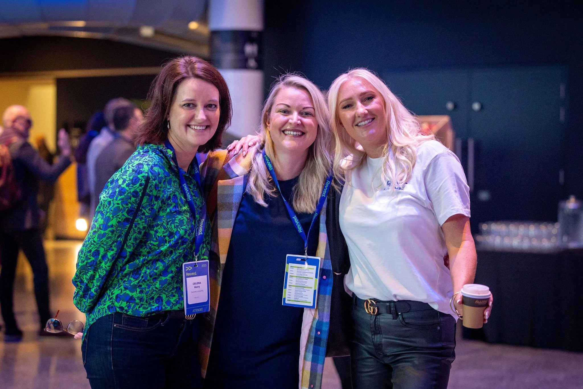 Three smiling women standing close together at an indoor event, wearing conference badges, with others blurred in the background.