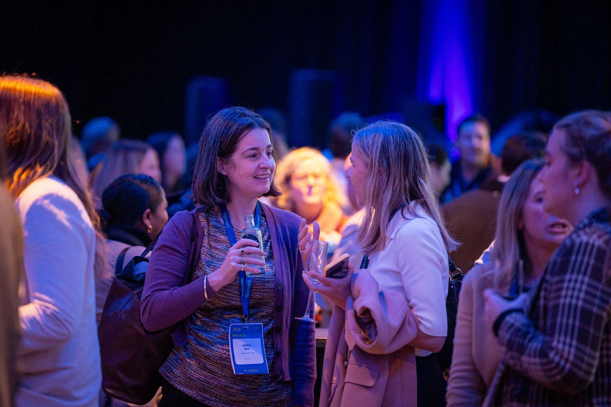 A group of people at a conference engaging in conversation, with a woman in the center holding a drink, wearing a name badge and purple jacket, surrounded by other attendees in a dimly lit, vibrant setting.