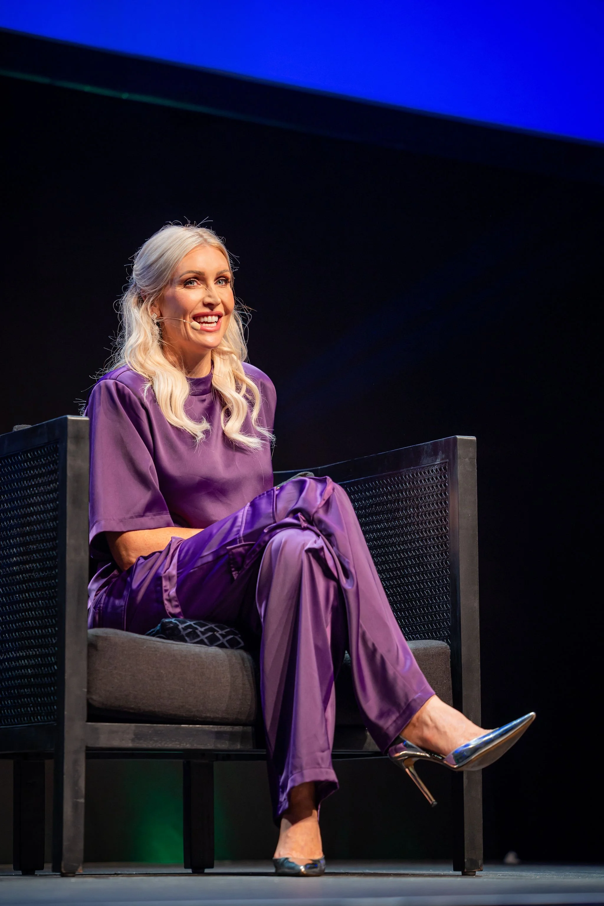 A woman with long blonde hair, wearing a purple dress, sitting on a dark-colored chair on a stage with a dark background, talking into a headset microphone and smiling.
