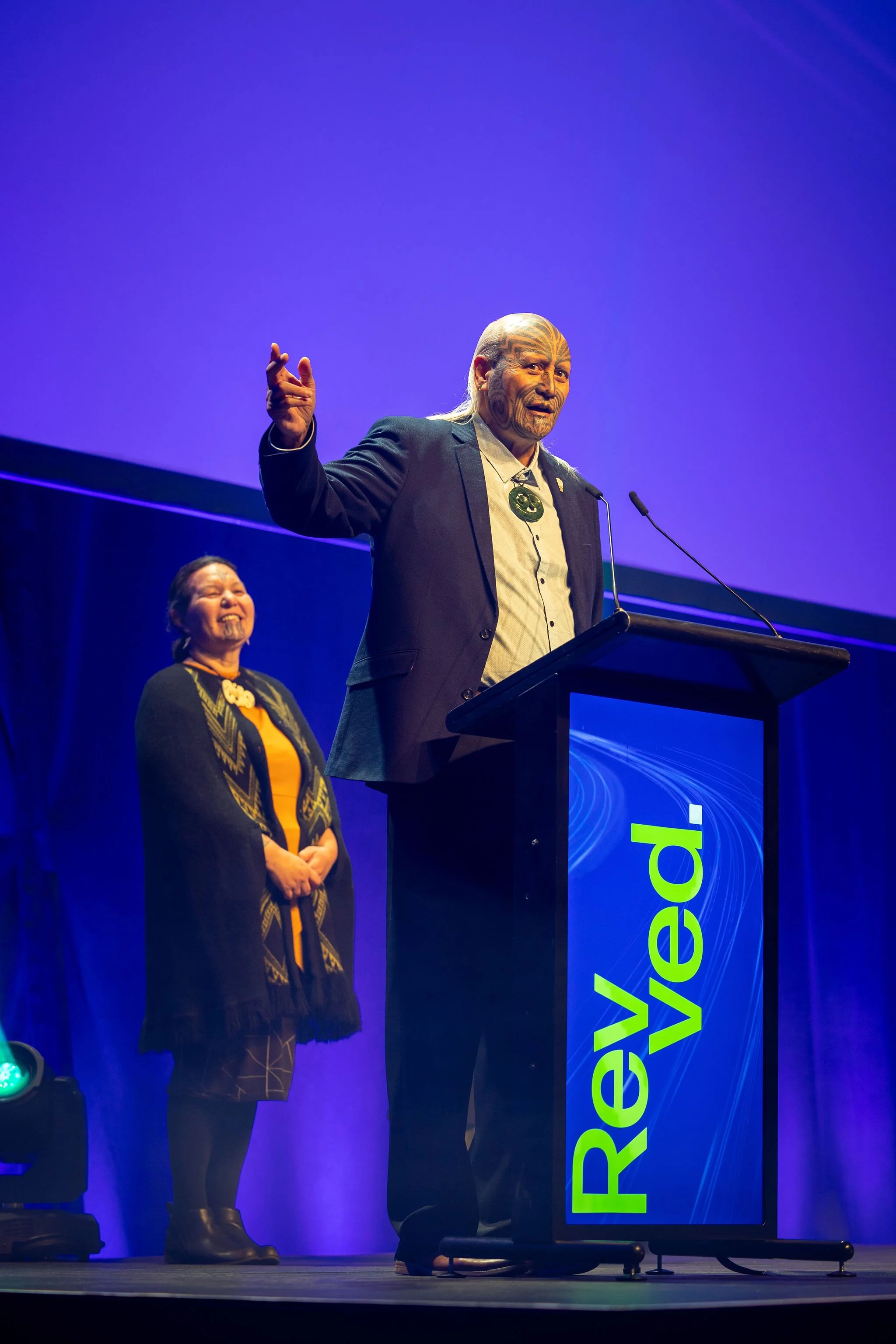 A man with tattoos speaking at a podium at Revved, with a woman standing behind him on stage. The stage is lit with purple and blue lighting, and there is a large screen with partially visible text.