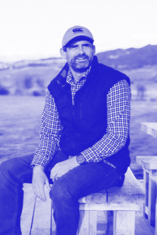 A man sitting outdoors on a wooden bench, wearing a baseball cap, glasses, a checkered shirt, and a vest, with a landscape and mountains in the background.