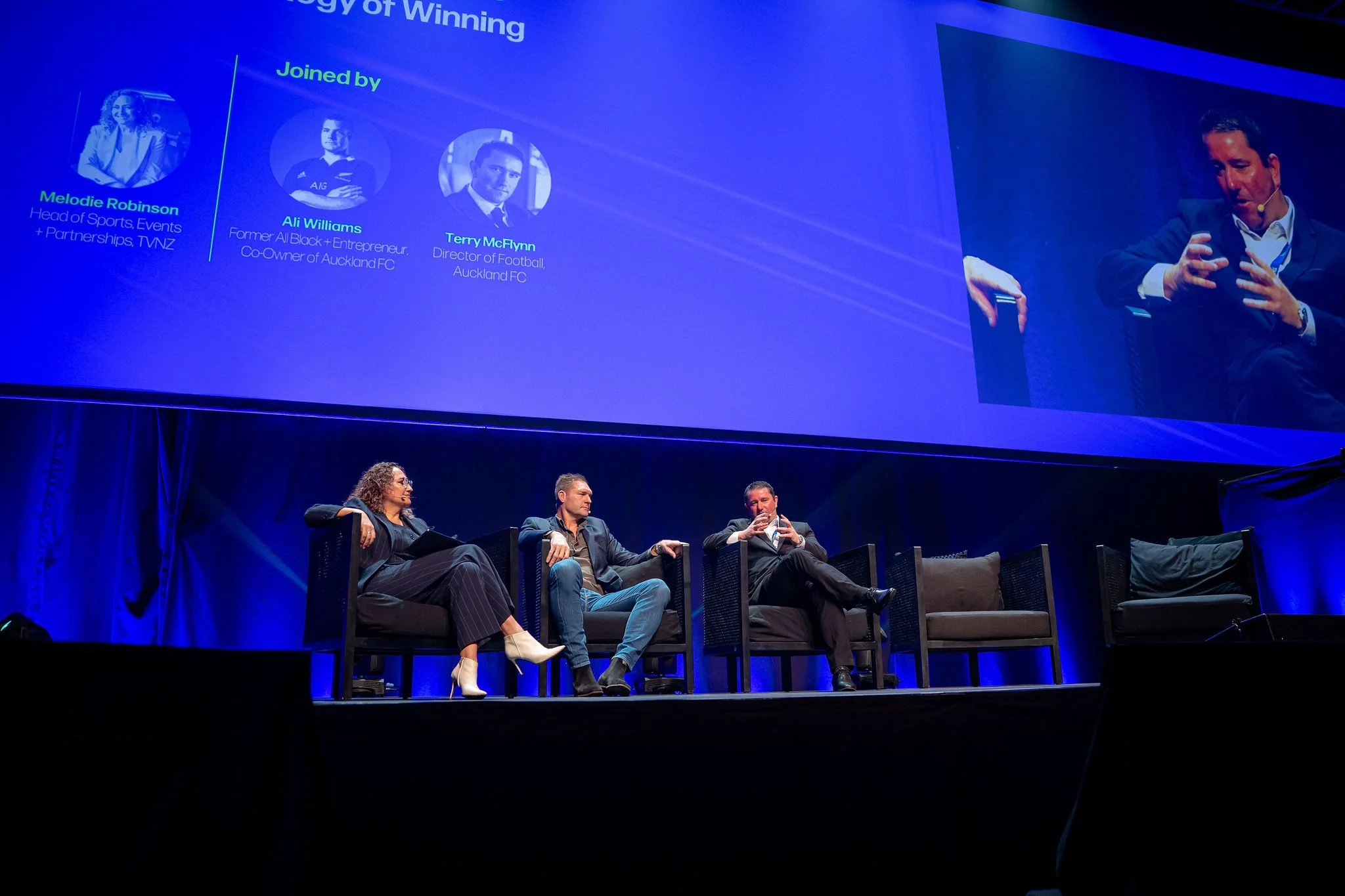 Revved panel discussion on stage with four people sitting in chairs, a large screen behind them displaying information about the panelists, including their names and titles, at a conference or event.