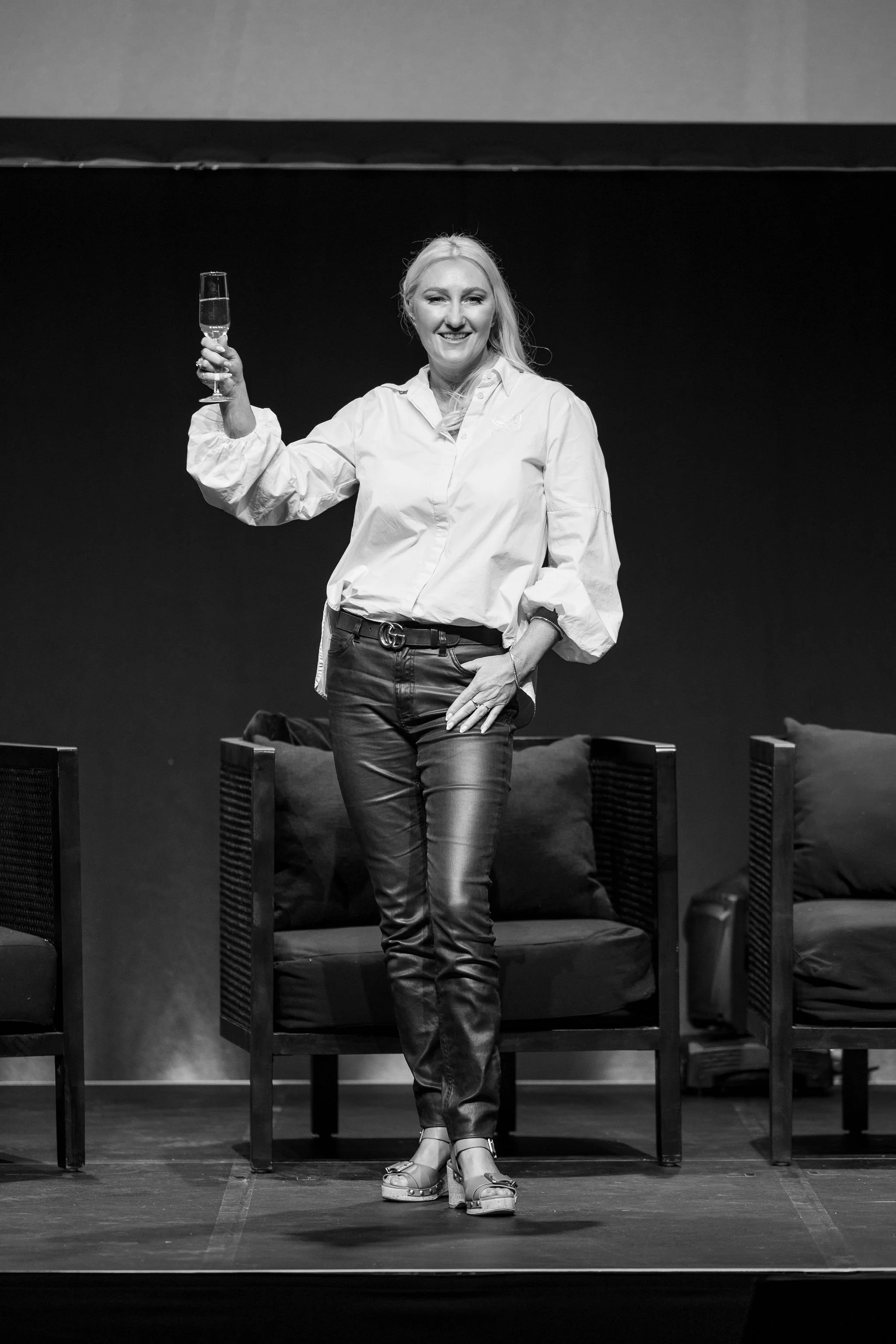 Black and white photo of a woman standing and smiling while holding a champagne glass in her right hand. She is wearing a white blouse, dark pants with a belt, and wedge sandals. The background has chairs with cushions.