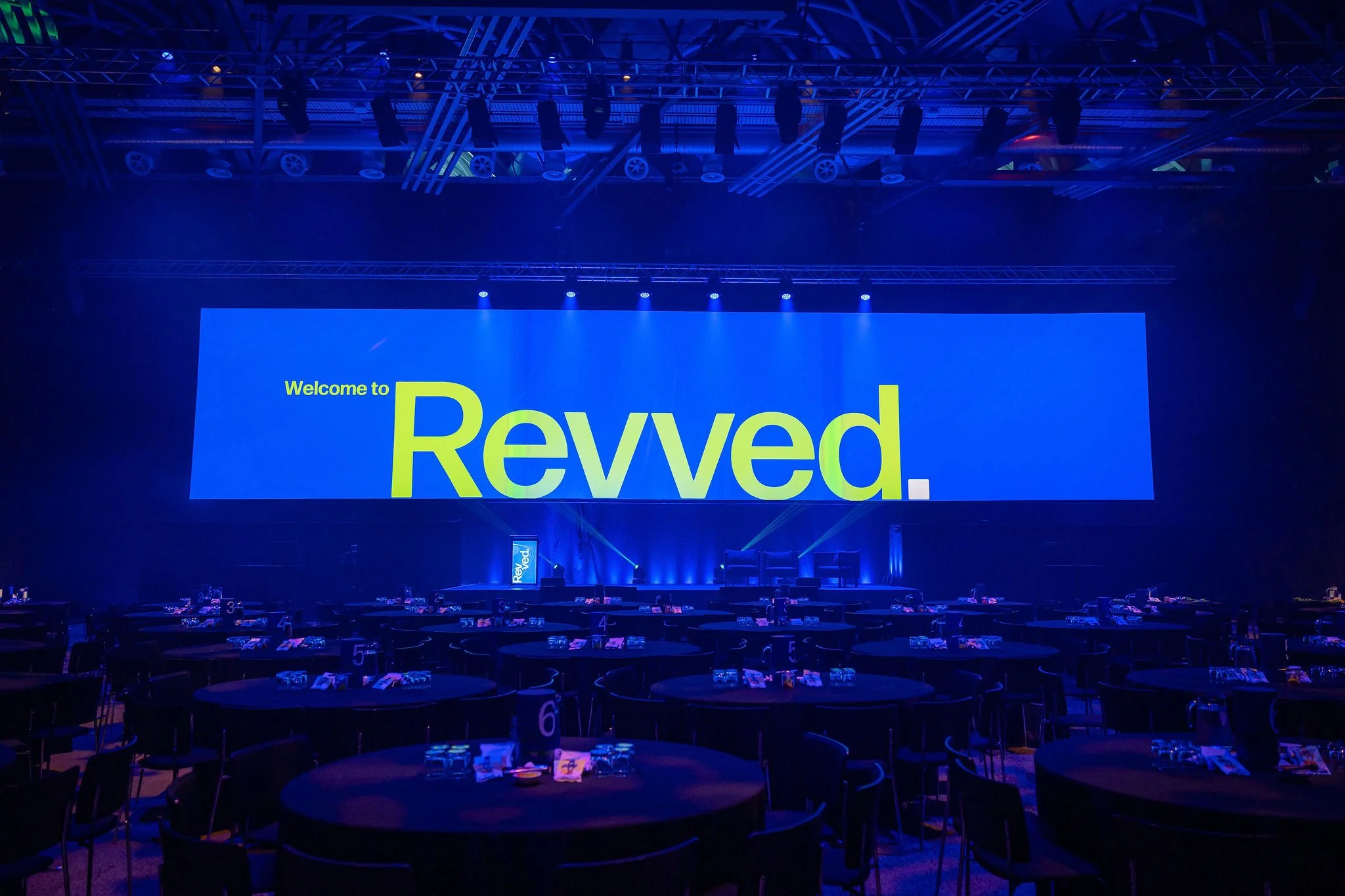 A large event hall with round tables set for a conference or banquet, dimly lit with blue lighting. A big screen displays the words "Welcome to Revved." hanging from the ceiling above the stage and tables.