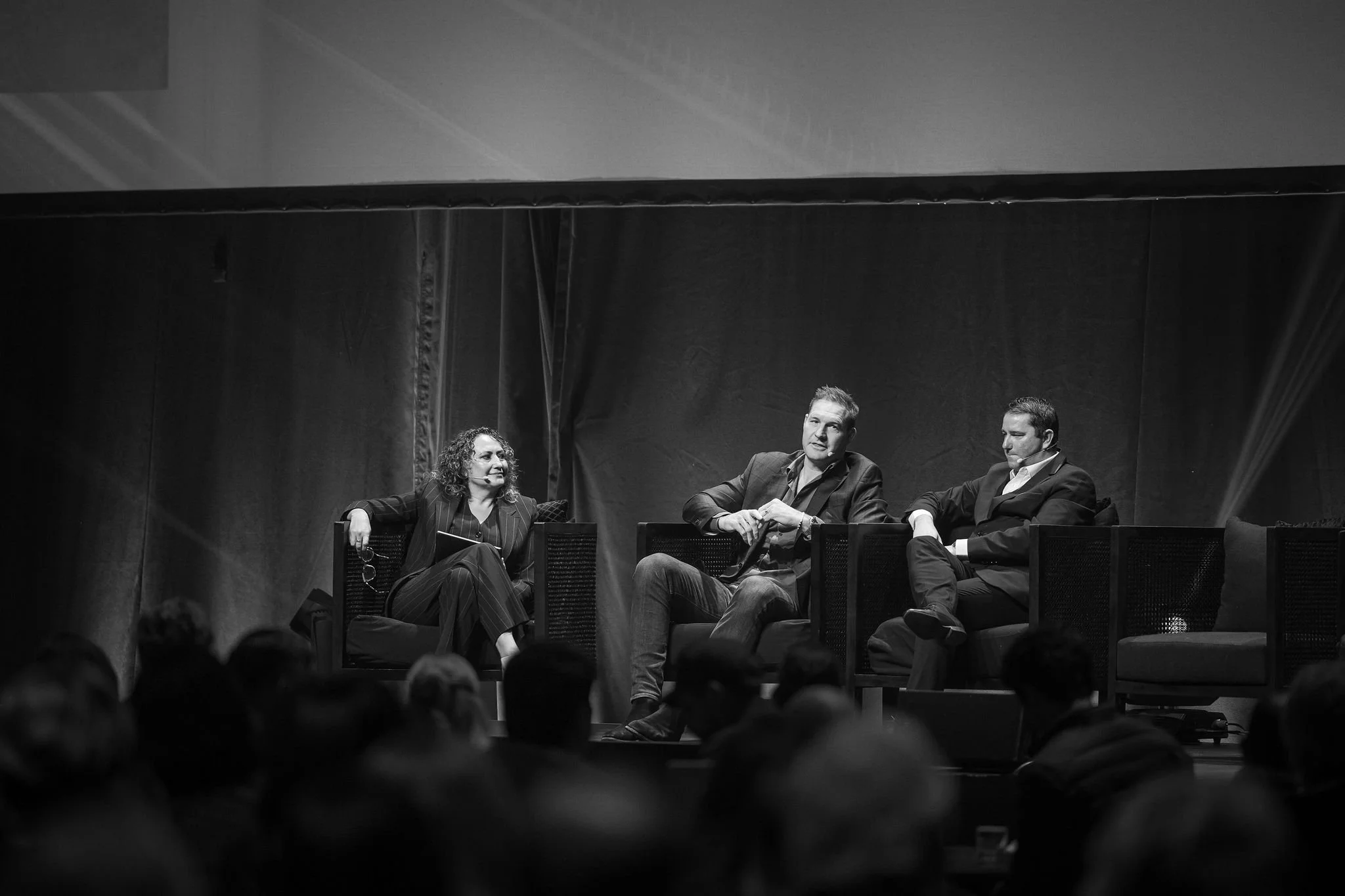 Black and white photograph of three people seated on a stage at Revved during a panel discussion, with an audience partially visible in the foreground.