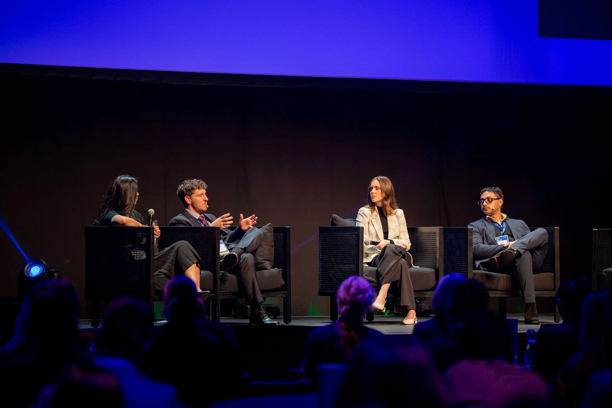 Four individuals seated on a stage in a panel discussion, with a dark background and blue lighting, audience visible at the bottom.