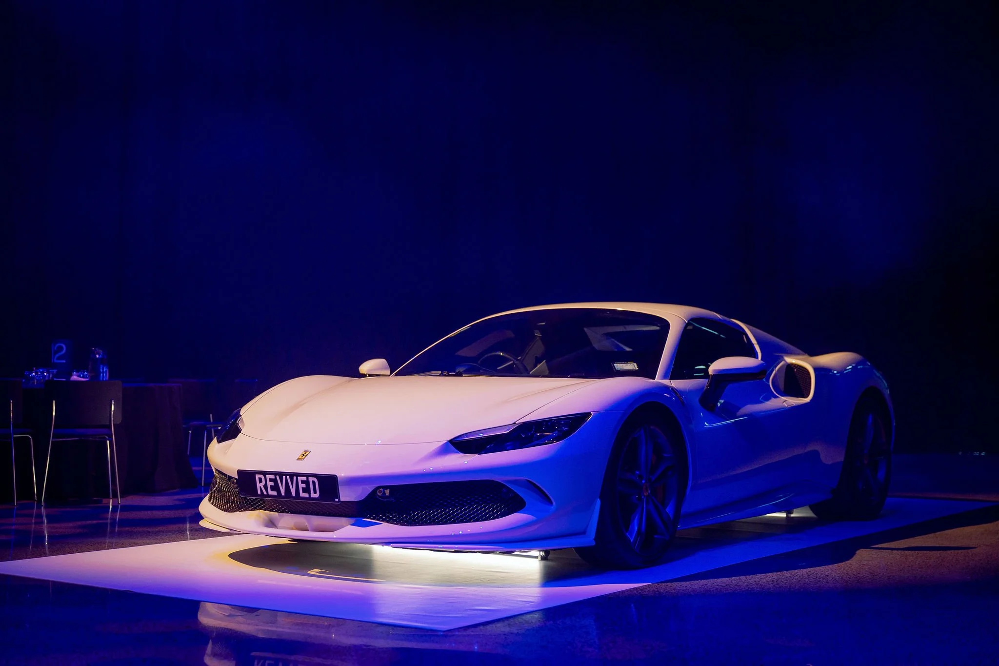 A white sports car with a Revspeed license plate displayed on a circular platform at an indoor event, illuminated by blue lighting.