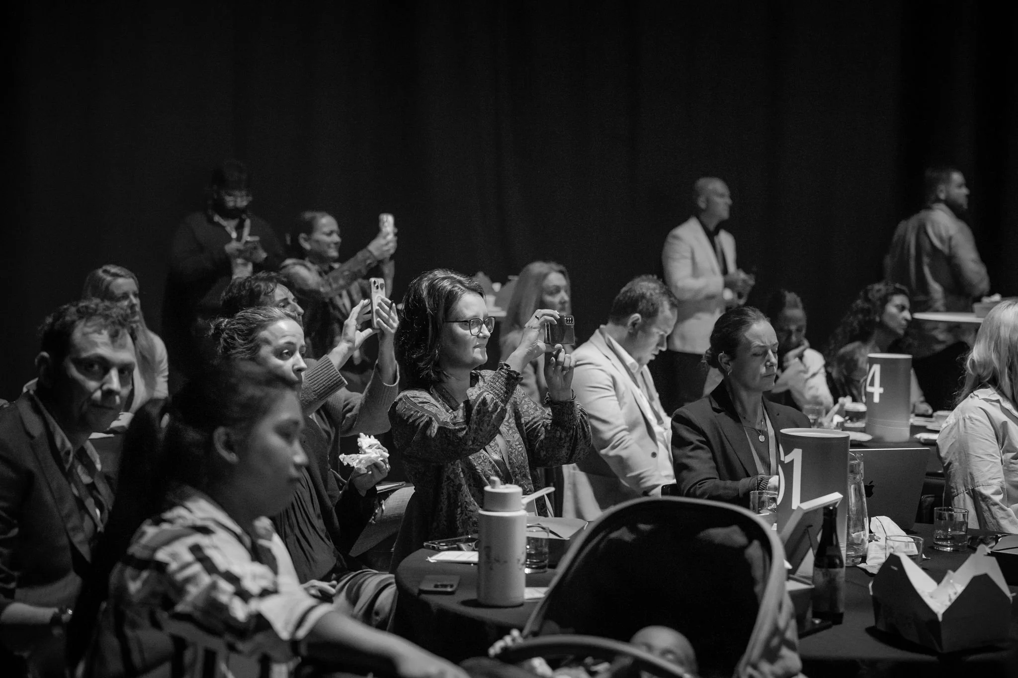 Audience at Revved conference, some taking photos or videos, some looking ahead, with tables, cups, and papers in front of them.