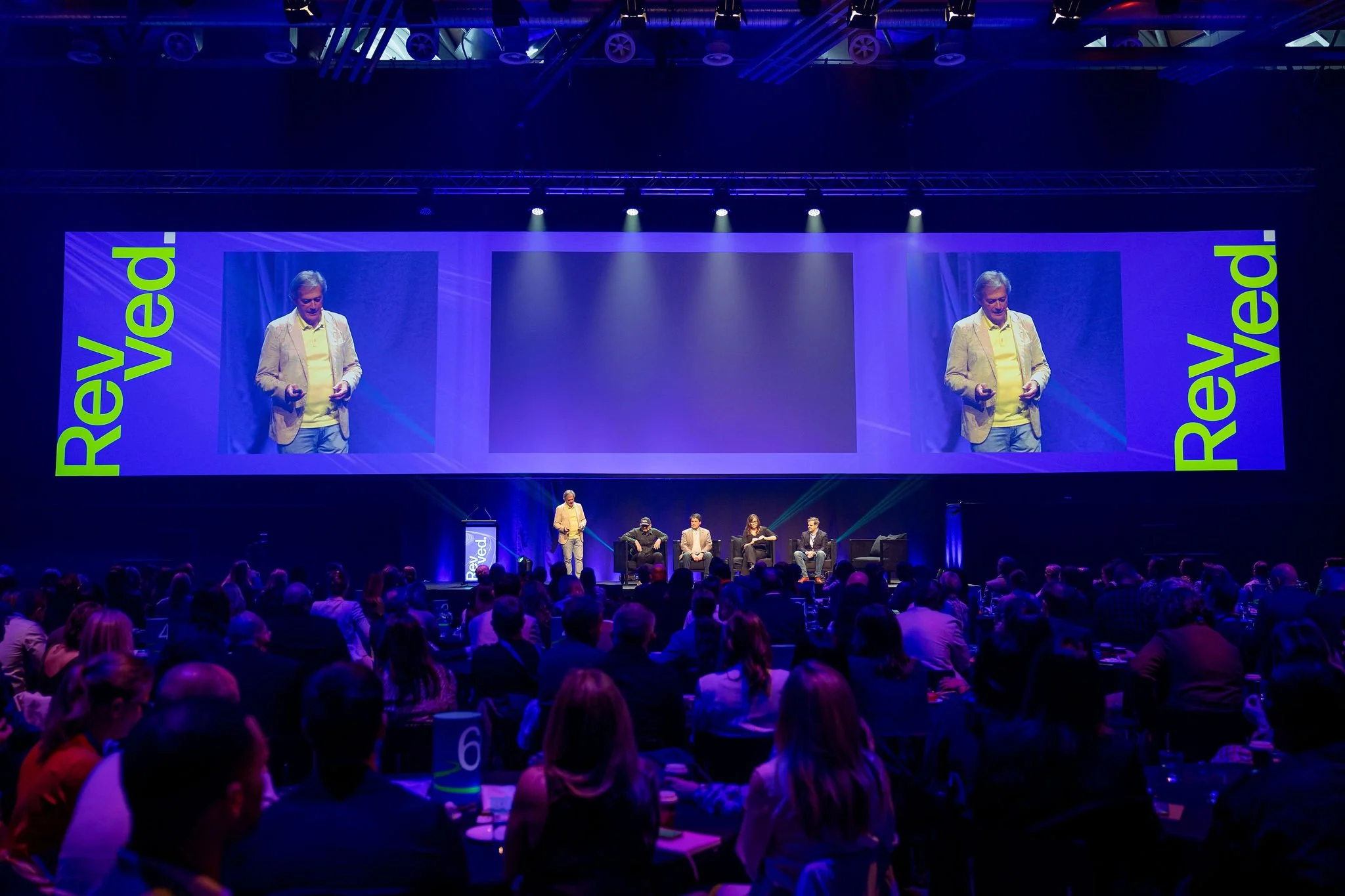 A speaker on stage at Revved. Multiple panelists are seated on stage, and the audience is seated at round tables.