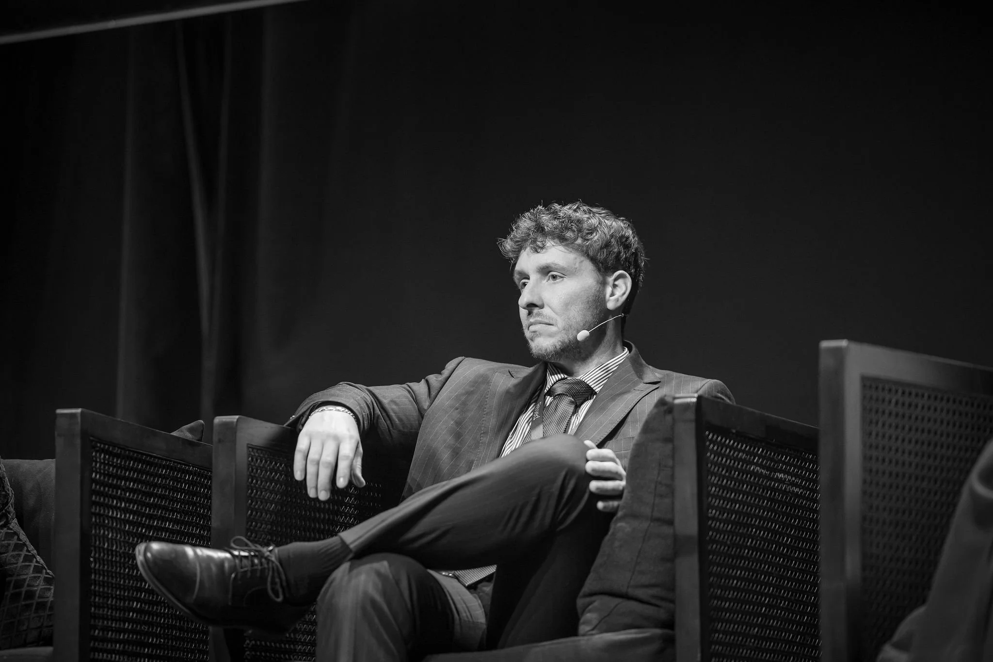 A man with curly hair and a mustache wearing a suit and tie, sitting on a chair with his arm resting on the armrest, in a black and white photo, speaking into a microphone, with a dark background.
