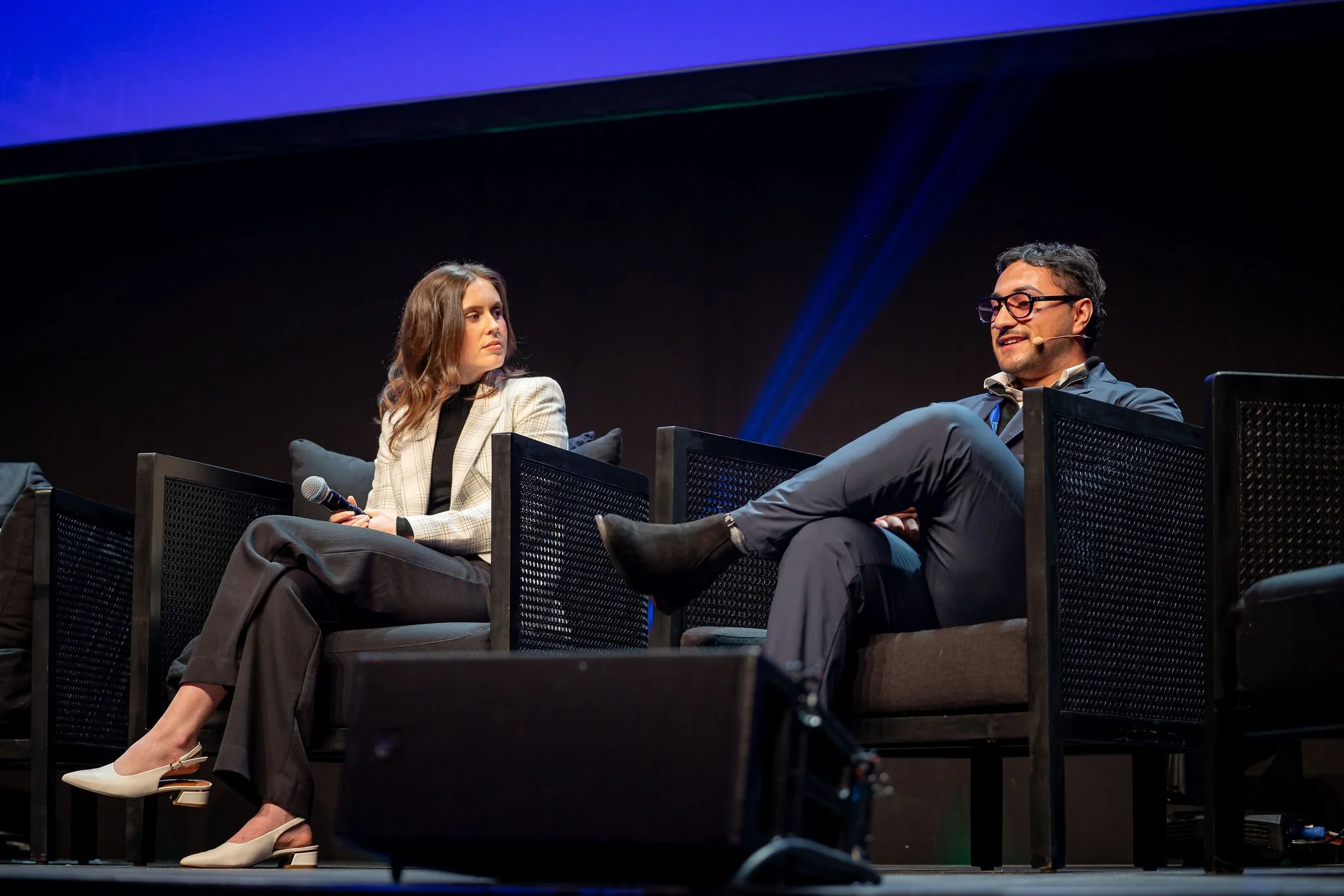 A man and a woman sitting on stage during a panel discussion, with the woman holding a microphone and the man speaking, both dressed in professional attire.
