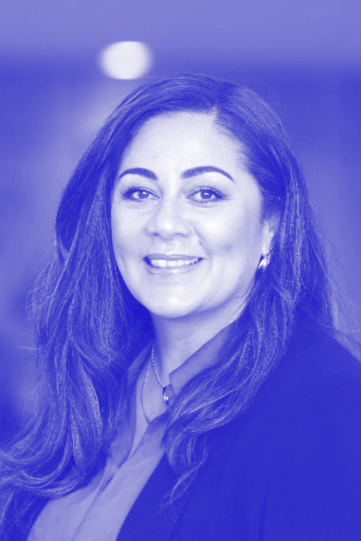 A woman with long dark hair smiling, wearing a blazer and a necklace, in a professional portrait setting, with a blurred background and a blue color overlay.