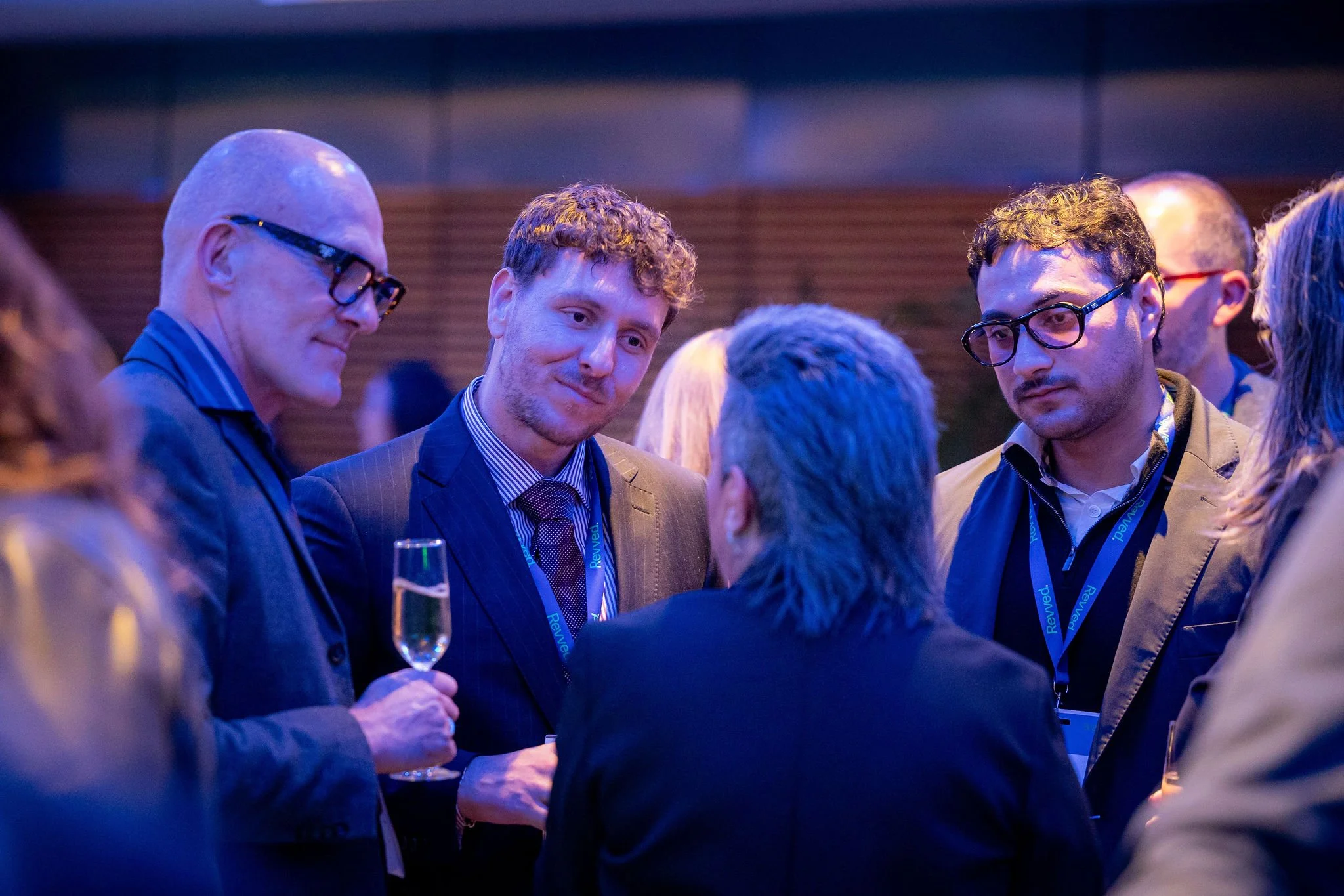 A group of men in business attire and sashes with blue ribbons are having a conversation at an event, with one holding a champagne flute.