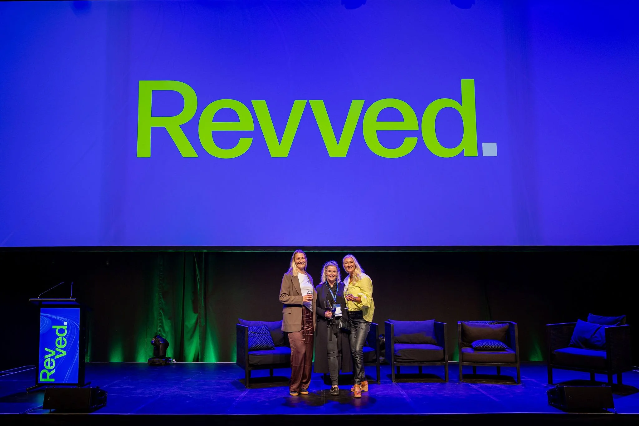 People standing on a stage at a conference, with a large screen behind them displaying the word "Revoked" in green text. There are three women in the photo, holding drinks and smiling.
