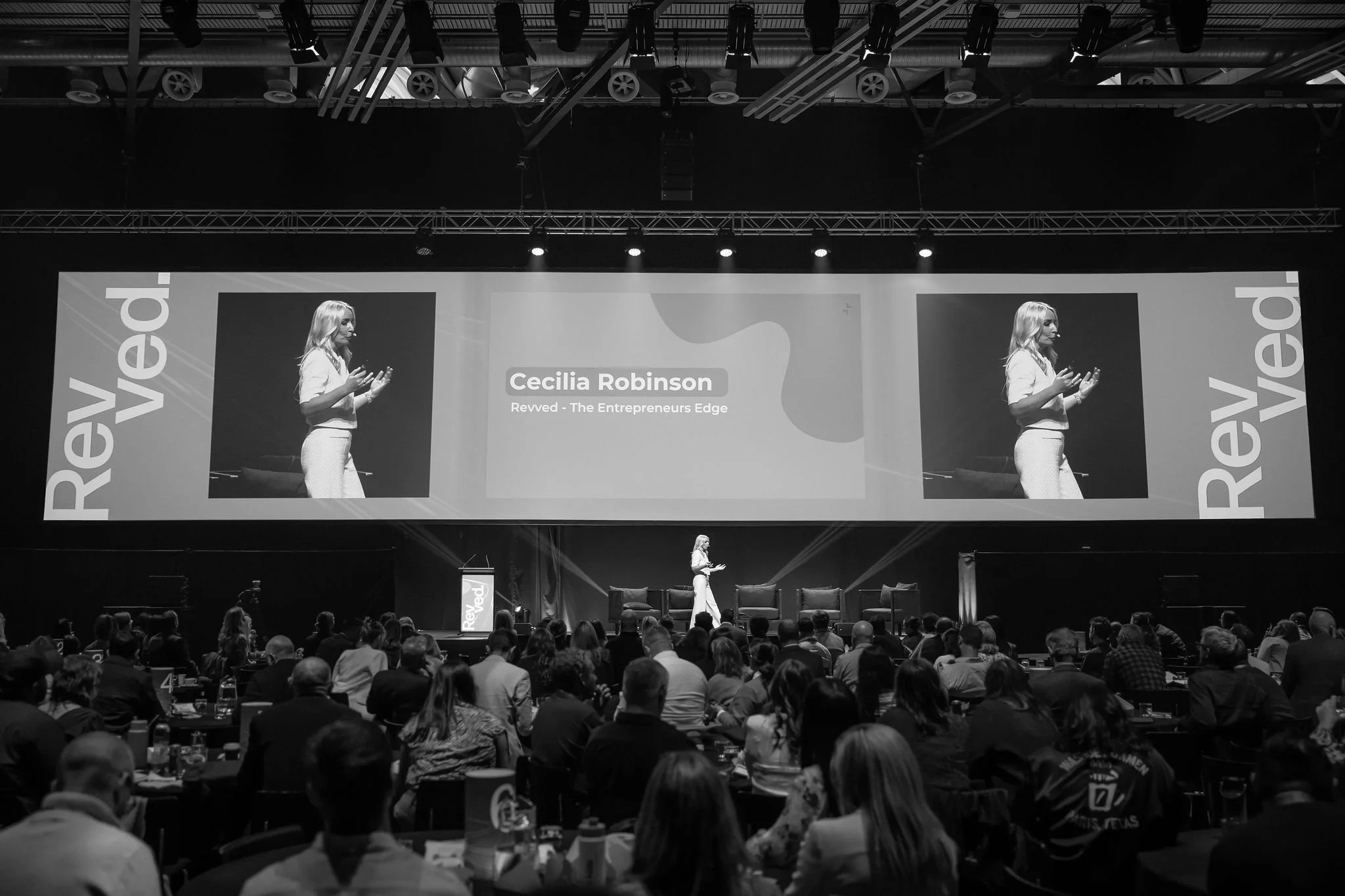 A woman is speaking on stage during a conference or seminar with a large audience. The stage has a screen behind her displaying her name, Cecilia Robinson, and the topic, Revved - The Entrepreneurs Edge. The conference appears to be called 'Reved,' a
