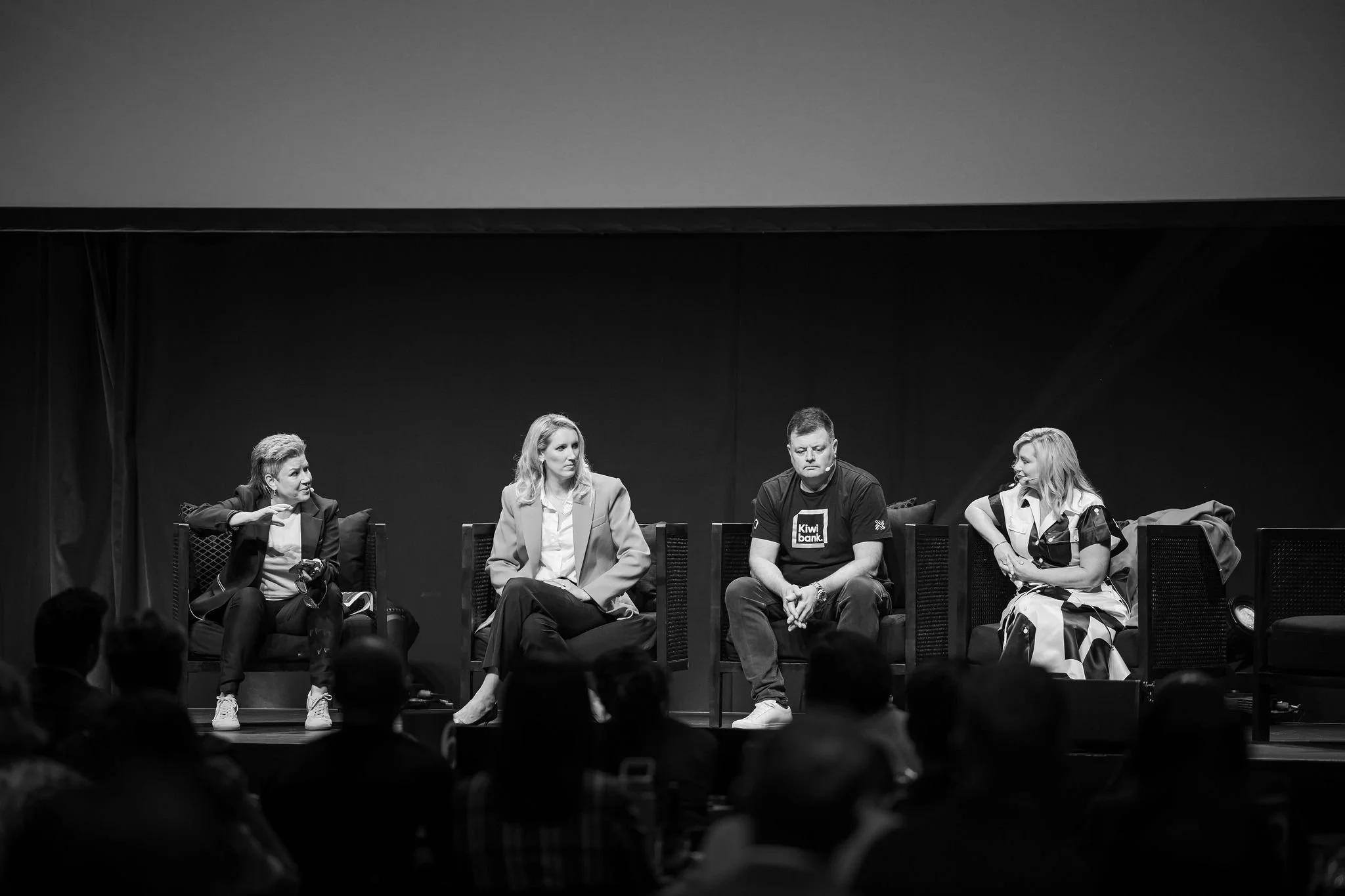 Black and white photo of four people sitting on stage during a panel or discussion, with an audience in front. The background is dark and plain.