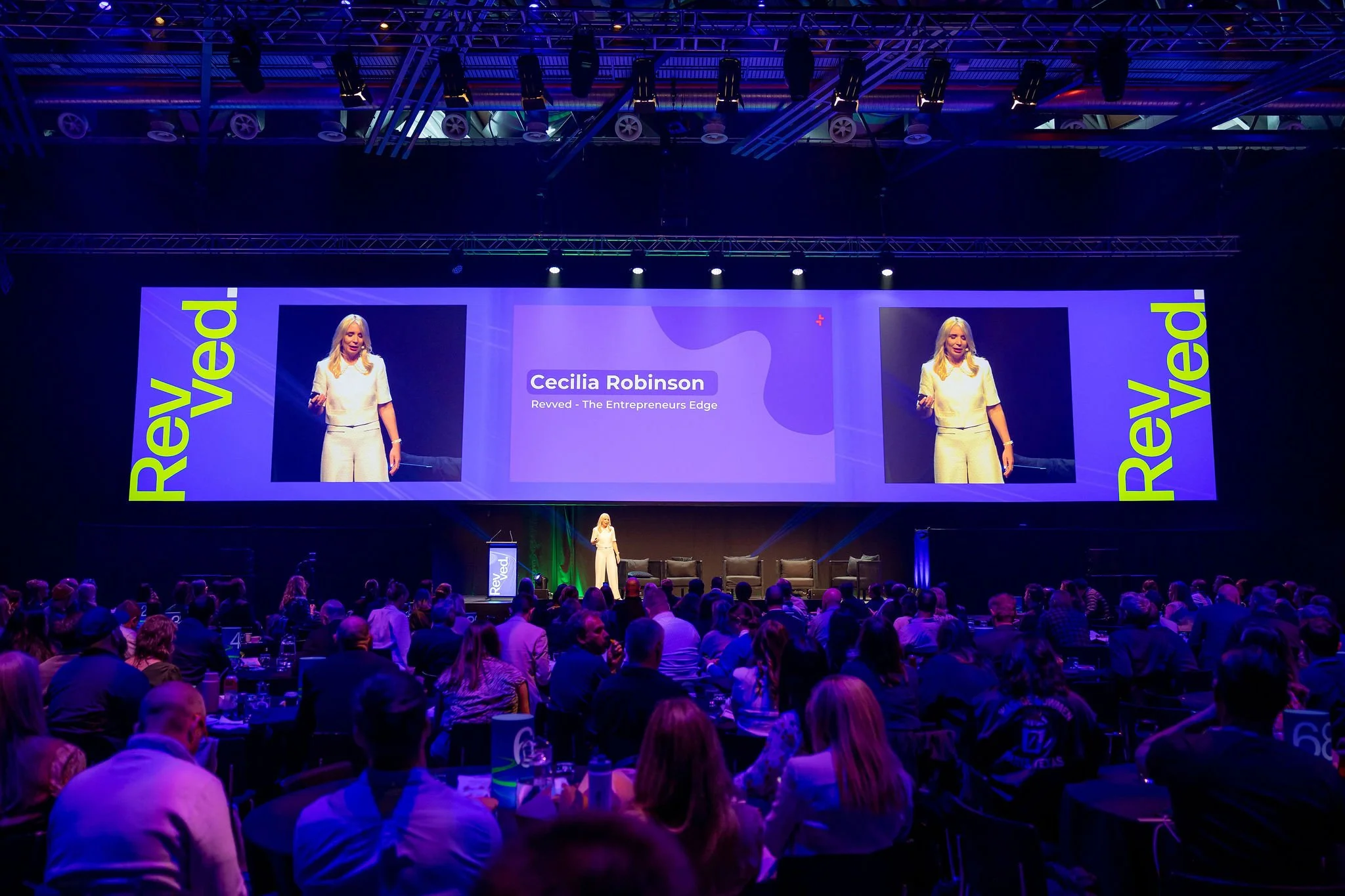 Cecilia Robinson standing on stage at conference, with large screen behind her displaying her name and title, audience seated in front.