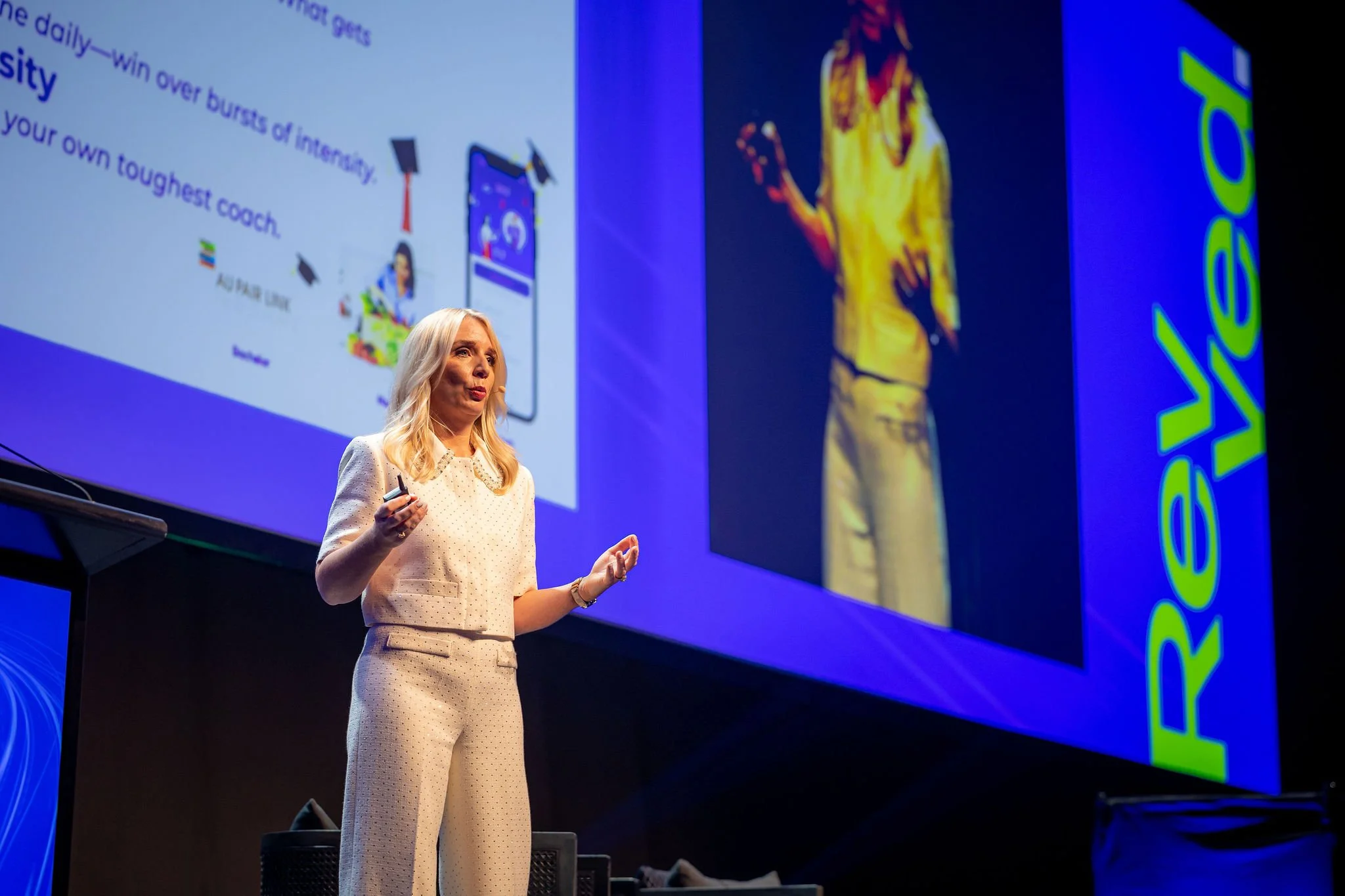 A woman with blonde hair giving a presentation on stage, holding a remote, with a slide projected behind her.