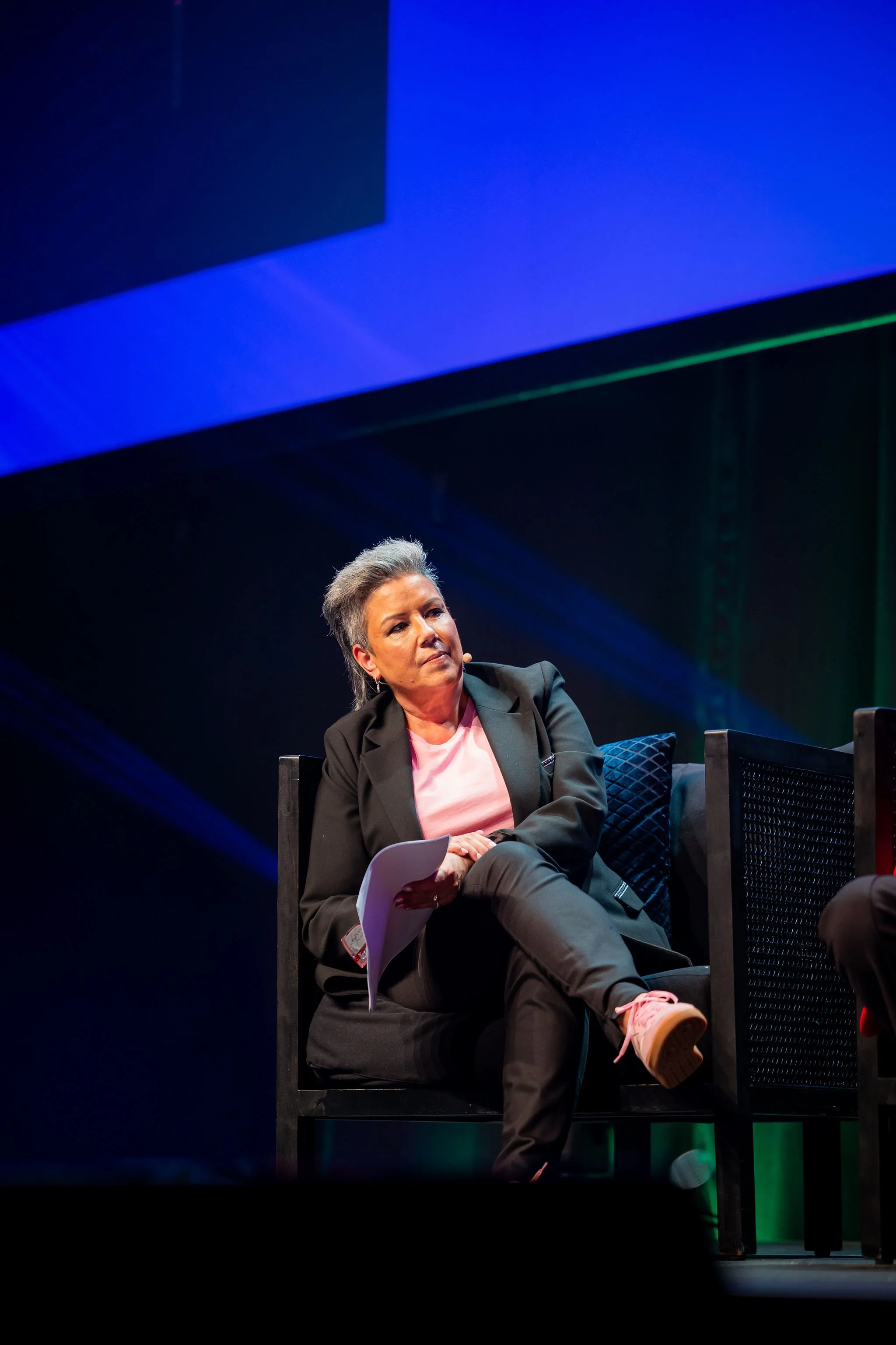 A woman with short gray hair, wearing a black blazer and pink sneakers, sitting on a black chair on a stage with blue and green lighting, holding papers.