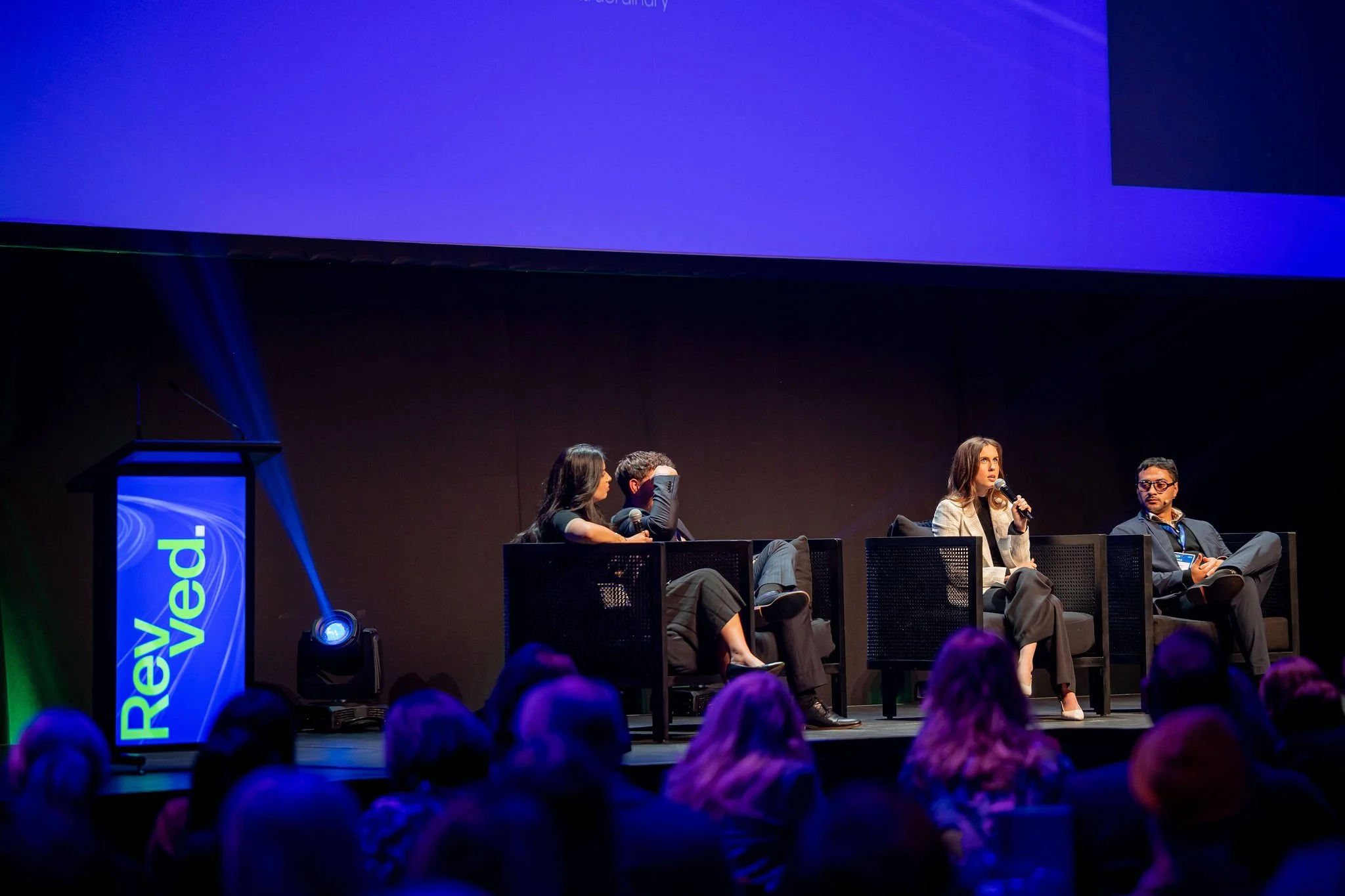 Panel discussion at a conference with four people seated on stage, microphone in hand, and a large blue screen overhead.