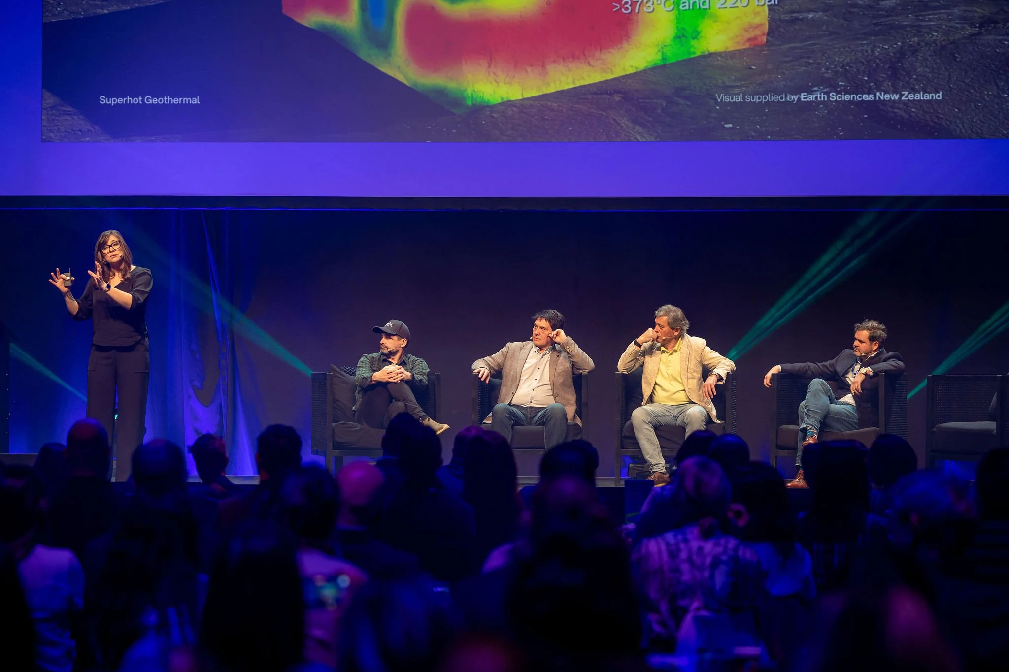 A woman in black attire presenting on stage at Revved with four seated men listening in front of an large presentation screen displaying a geothermal diagram and text.